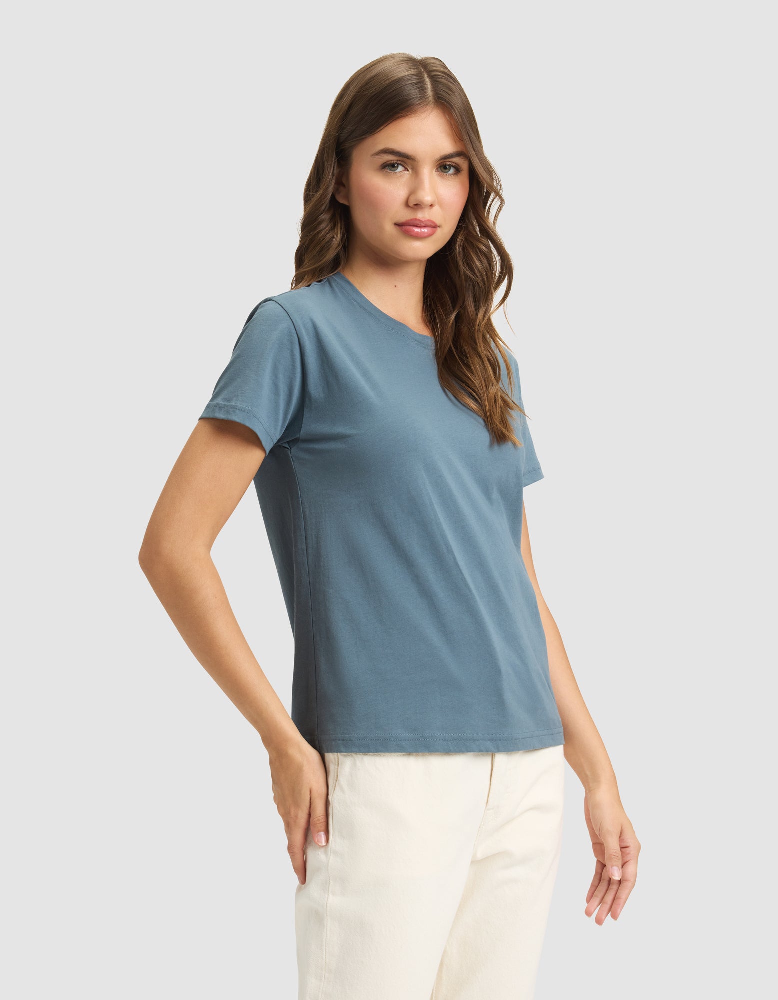 A woman with long brown hair wears the Cozy Earth Women's Easy Tee in blue and off-white pants, standing against a plain light gray background. She faces the camera with a neutral expression. 