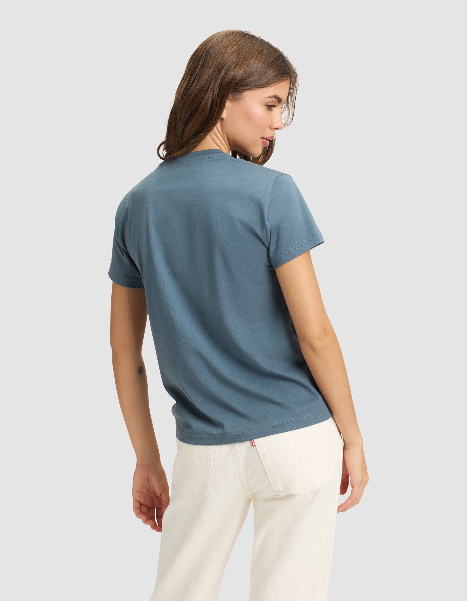 A woman with light brown hair models the Cozy Earth Women's Easy Tee in blue and off-white pants, standing with her back partially turned against a plain light gray background. 