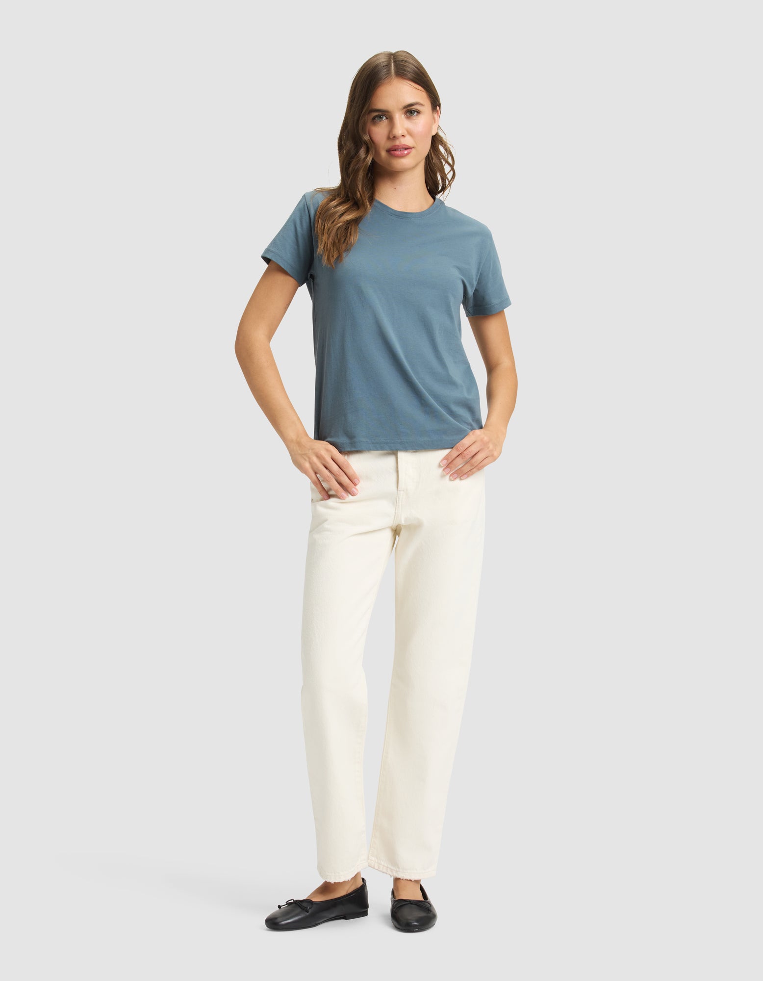A woman with long, wavy hair stands facing forward against a plain light gray background, wearing the Cozy Earth Women's Easy Tee in blue, off-white pants, and black slip-on shoes, with her hands resting on her hips. 