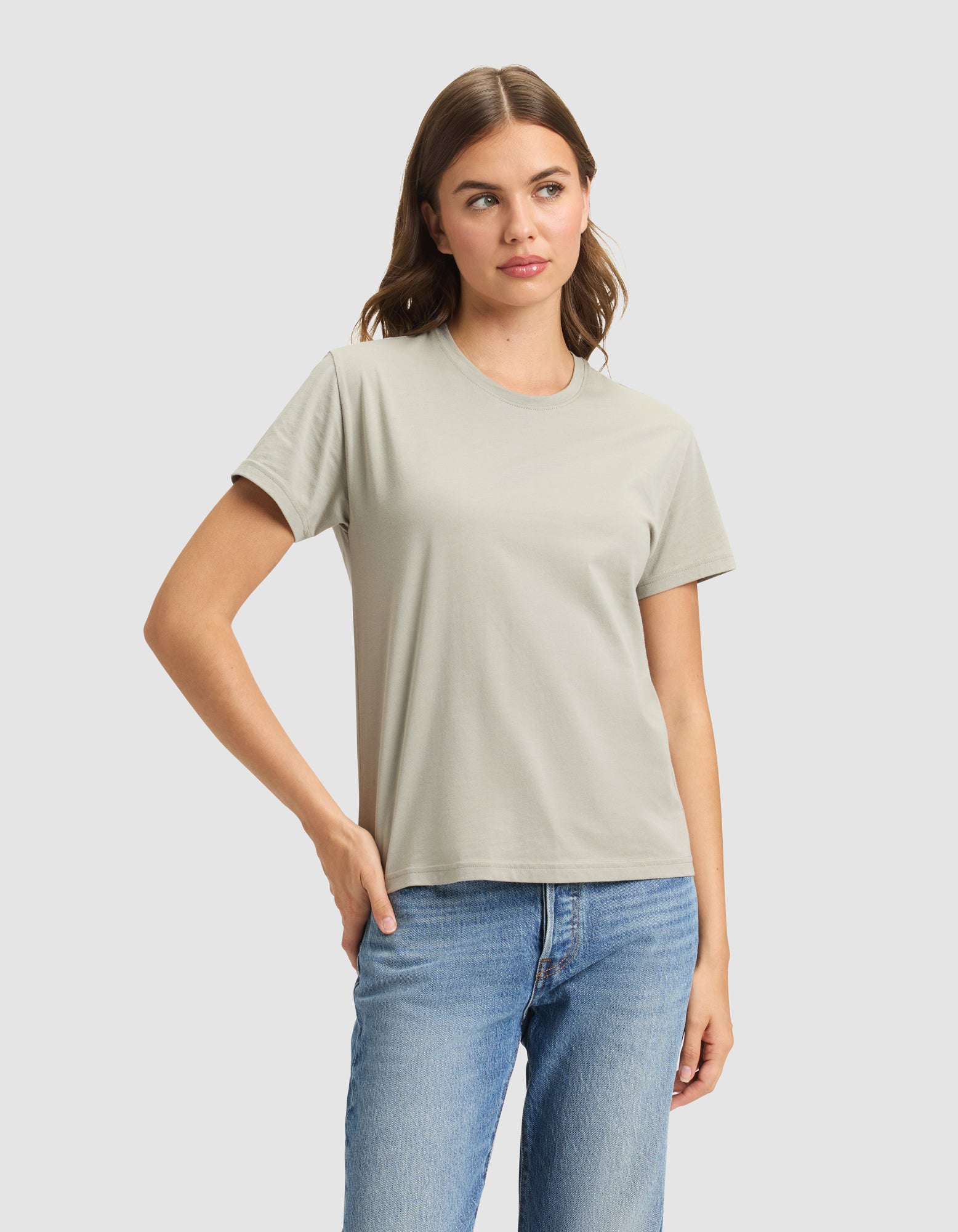 A woman wears the Cozy Earth Women's Easy Tee in light gray with blue jeans, standing against a plain light background, one hand on her hip and a neutral expression. |Color:Palm