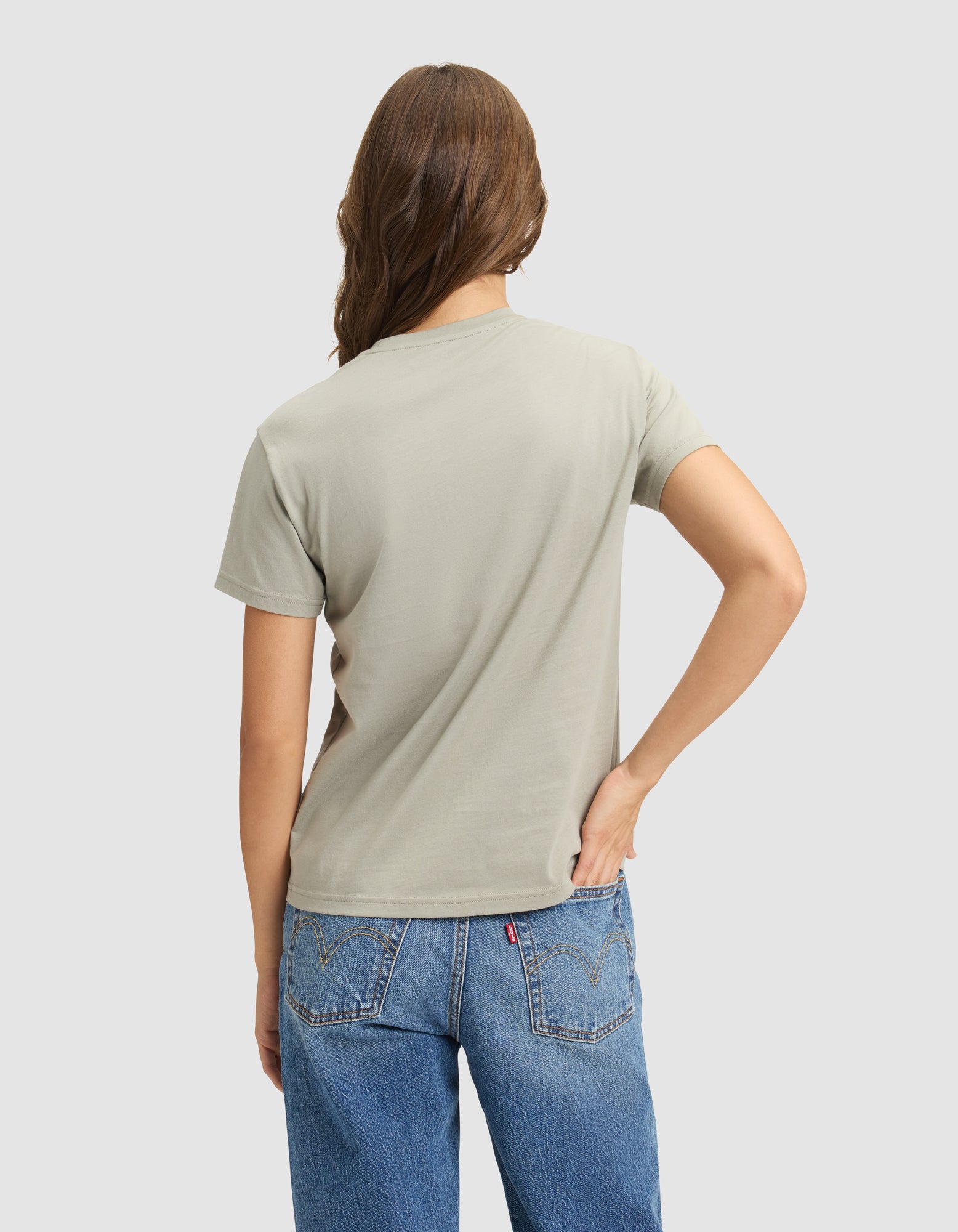 A woman with long brown hair wears the Cozy Earth Women's Easy Tee in light gray and blue jeans, standing with one hand on her hip, facing away from the camera against a plain light background. 