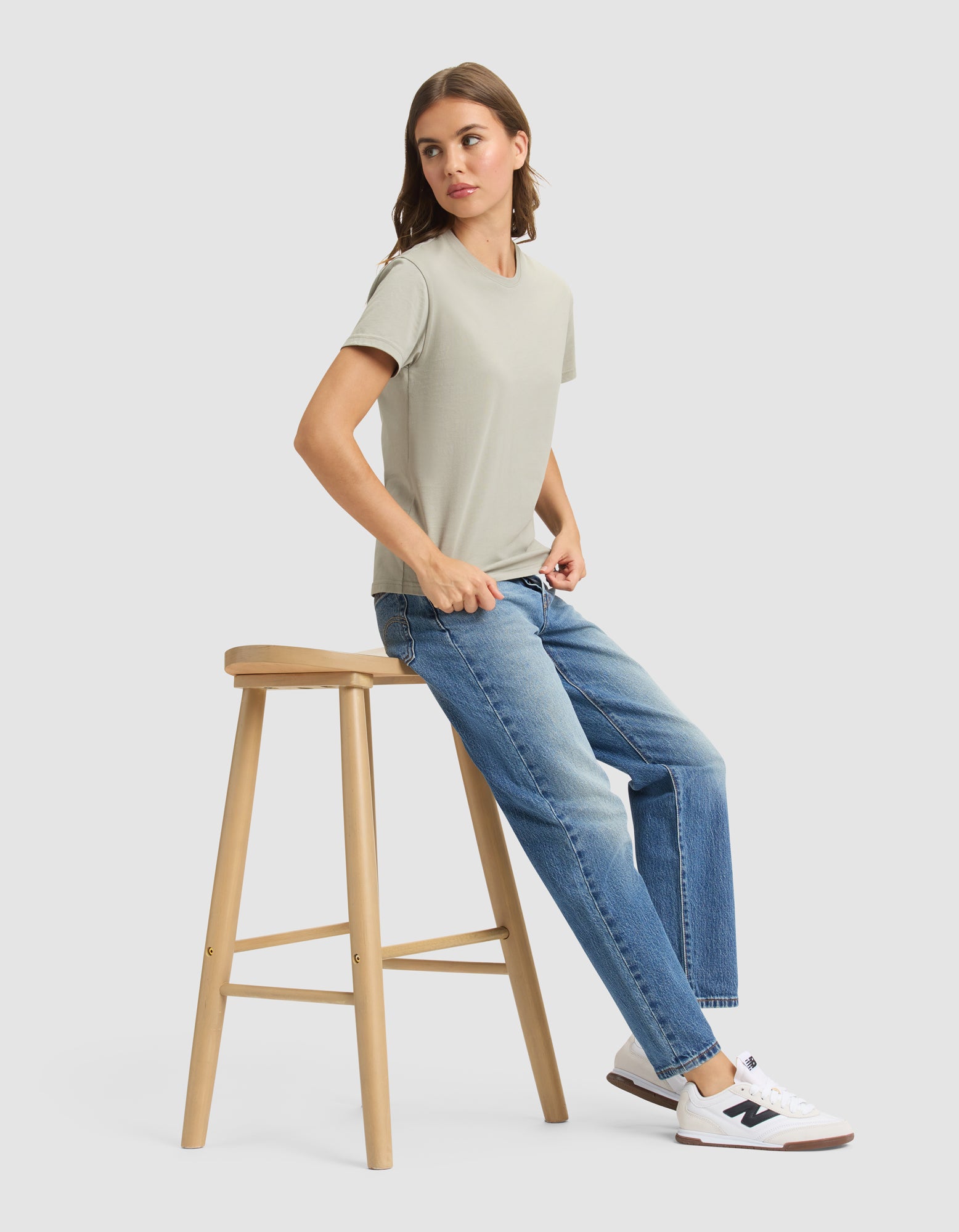 A woman wearing the Cozy Earth Women's Easy Tee in light green, blue jeans, and white sneakers sits sideways on a wooden stool against a plain light gray background. 