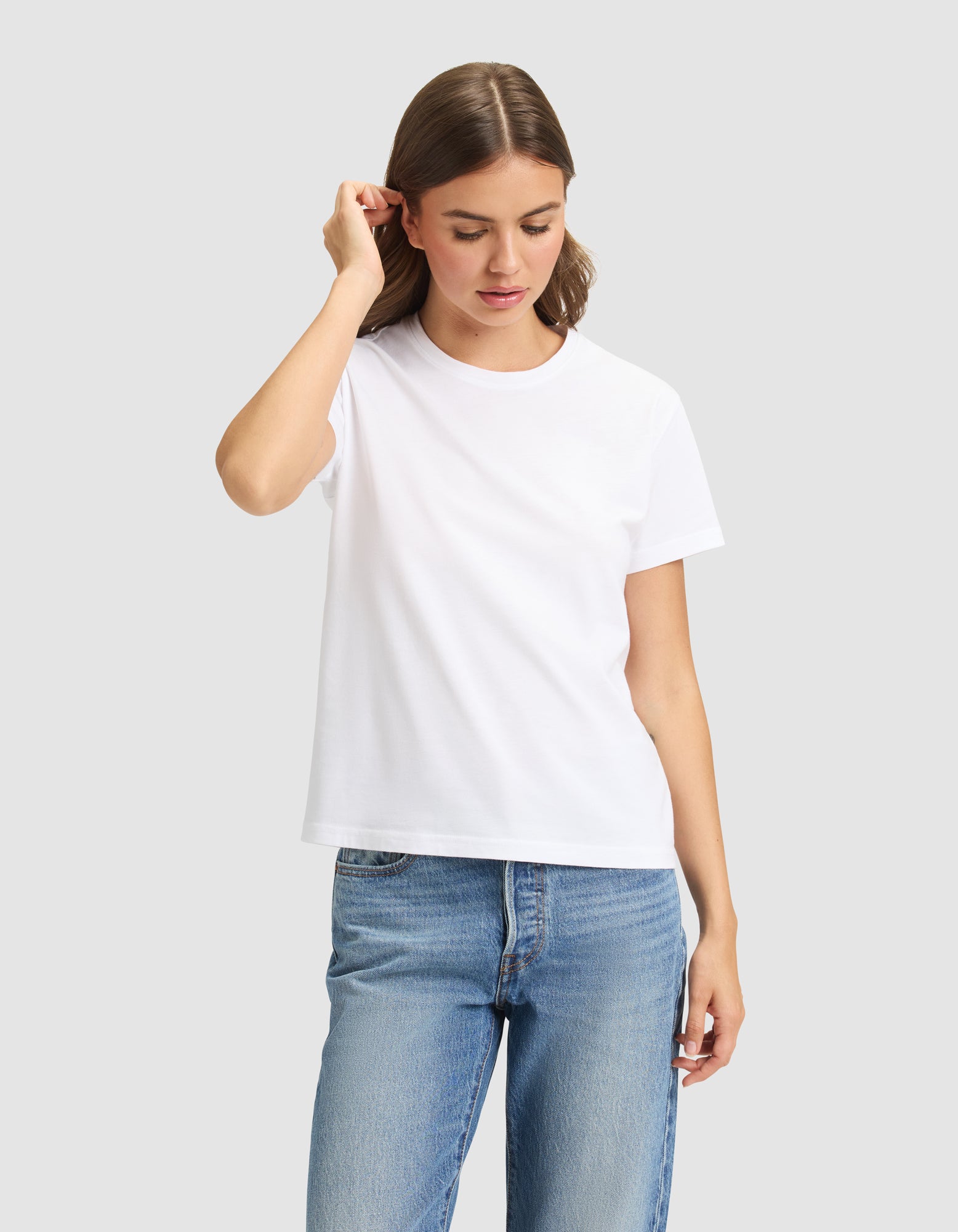 A woman with shoulder-length brown hair models the Cozy Earth Women's Easy Tee in white, paired with blue jeans, while standing against a light gray background and touching her hair. 