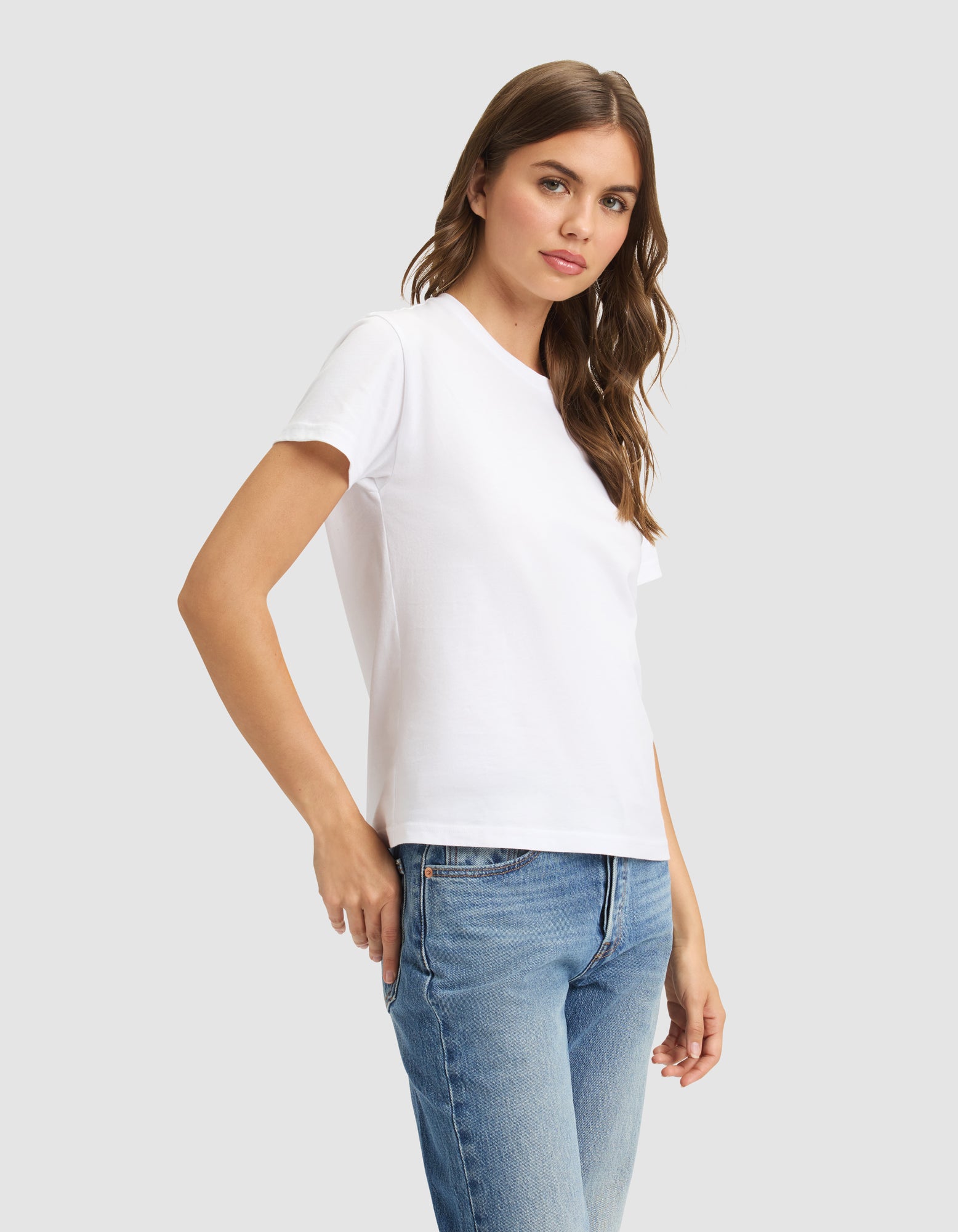 A woman with long brown hair models the Cozy Earth Women's Easy Tee in white with blue jeans against a light gray background, gazing at the camera with a neutral expression. 