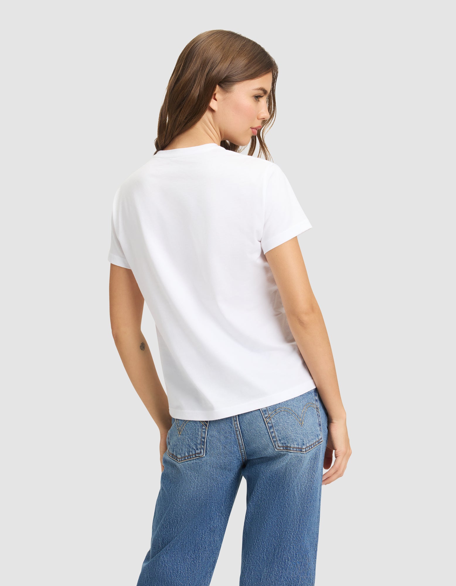 A woman with long brown hair stands facing away from the camera, wearing Cozy Earth's Women's Easy Tee and blue jeans against a light gray background. 
