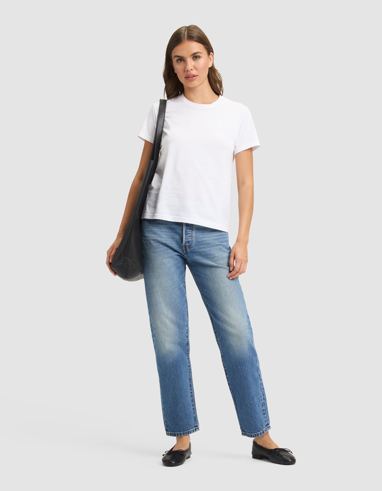 A woman stands against a plain background, wearing the Cozy Earth Women's Easy Tee in white, blue jeans, black flats, and carrying a black shoulder bag. 