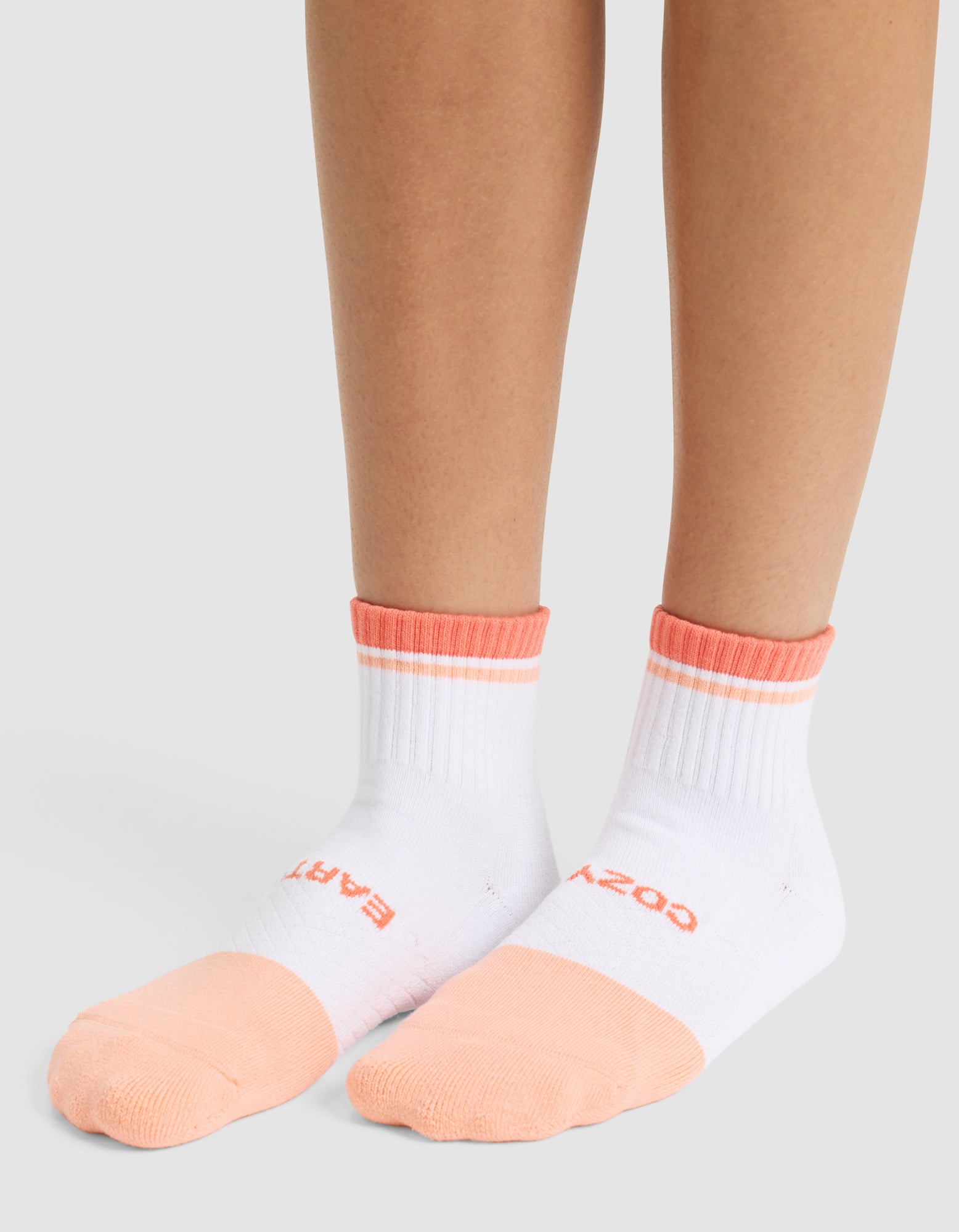 A person wears Cozy Earth Essential Quarter Socks in white with peach toes, heels, and cuffs. The socks feature text near the toes and are shown on bare legs against a light gray background. 