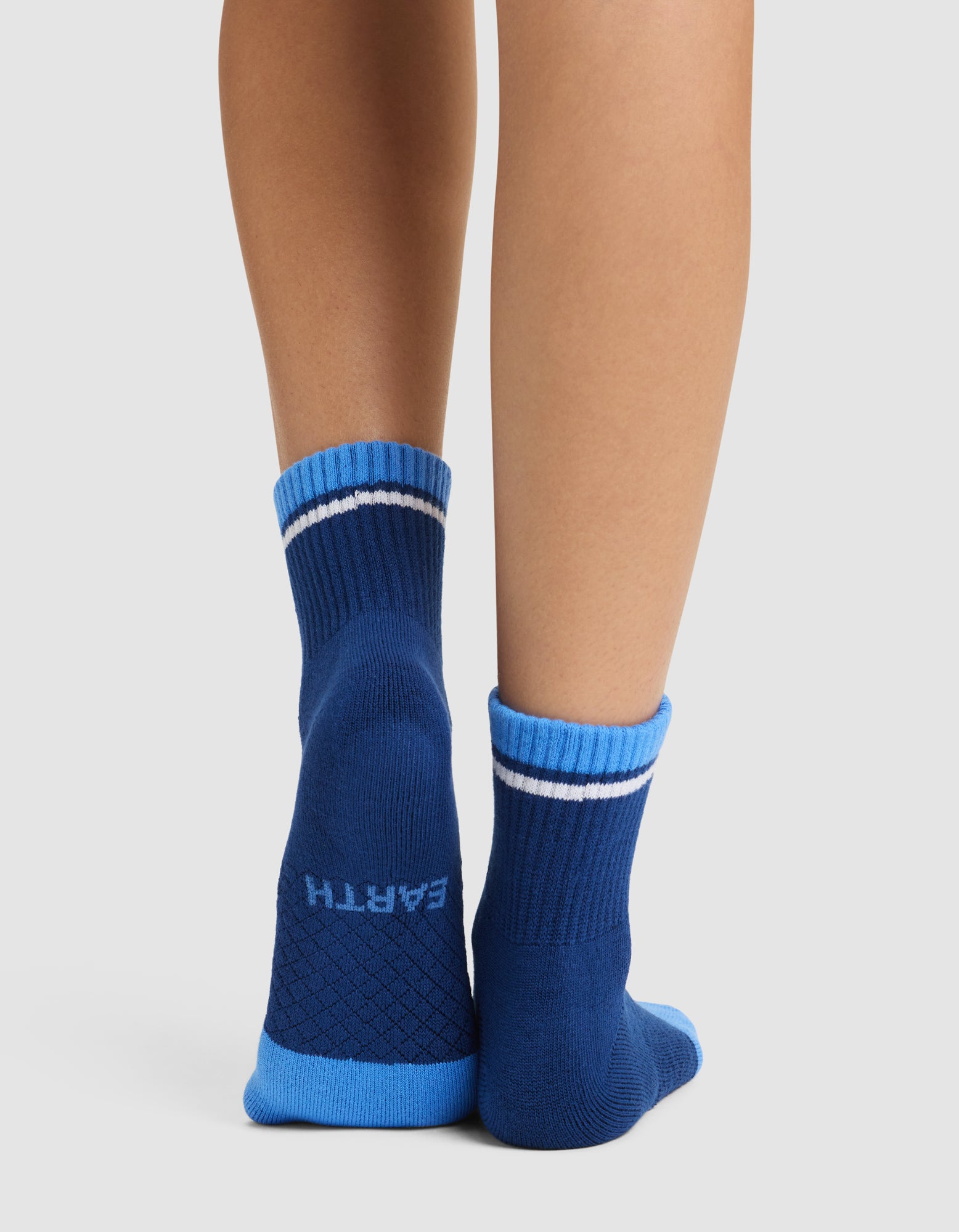 A person stands on tiptoe wearing Cozy Earth Essential Quarter Socks in blue with light blue toes, heels, and cuffs. "EARTH" is written in white on one sole. The plain background is light-colored. 