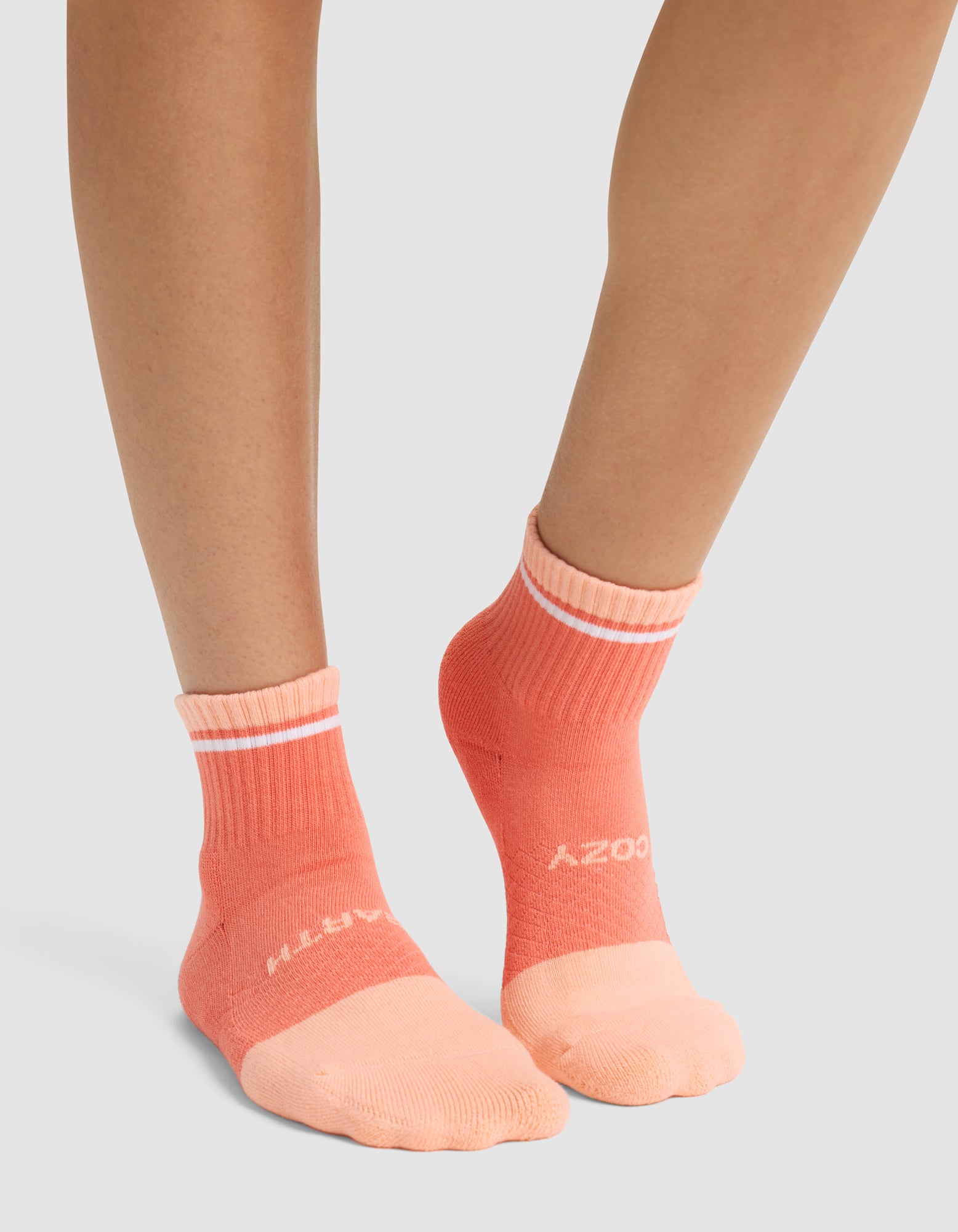 A person stands on a light gray surface wearing Cozy Earth's Essential Quarter Sock in peach and coral, featuring white top stripes and white text. 