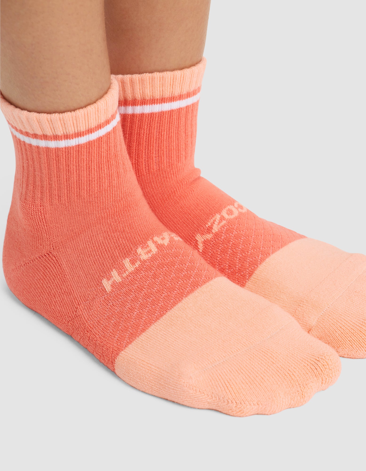 Two feet wear coral Essential Quarter Socks by Cozy Earth, featuring lighter peach toes and heels with a white stripe near the top. The plain light gray background highlights this stylish sock from the Cozy Earth 4-Pack set. 
