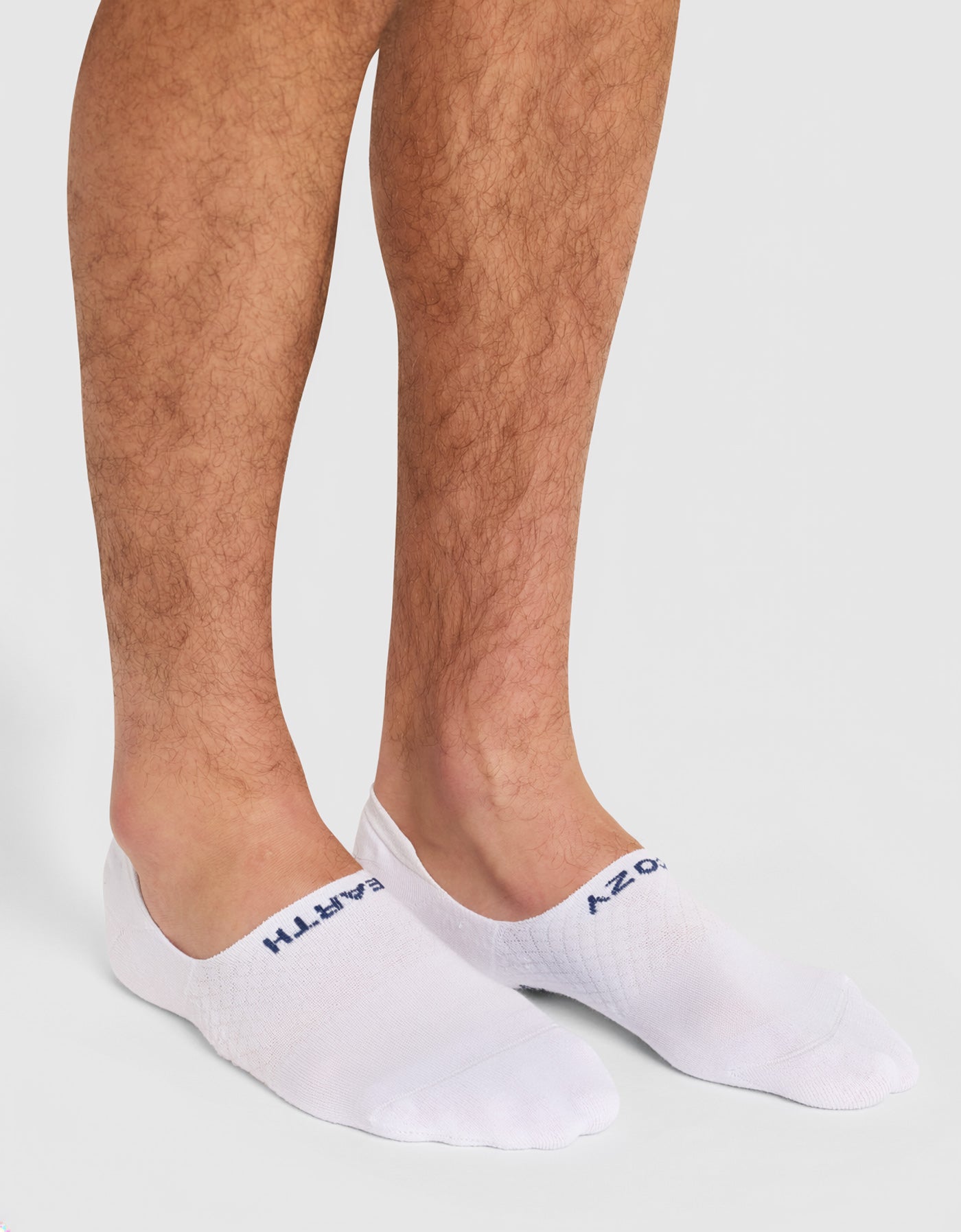 A person wears Cozy Earth Men's Essential No-Show Socks (white with blue text) while standing on a light gray surface; only their lower legs and feet are visible.