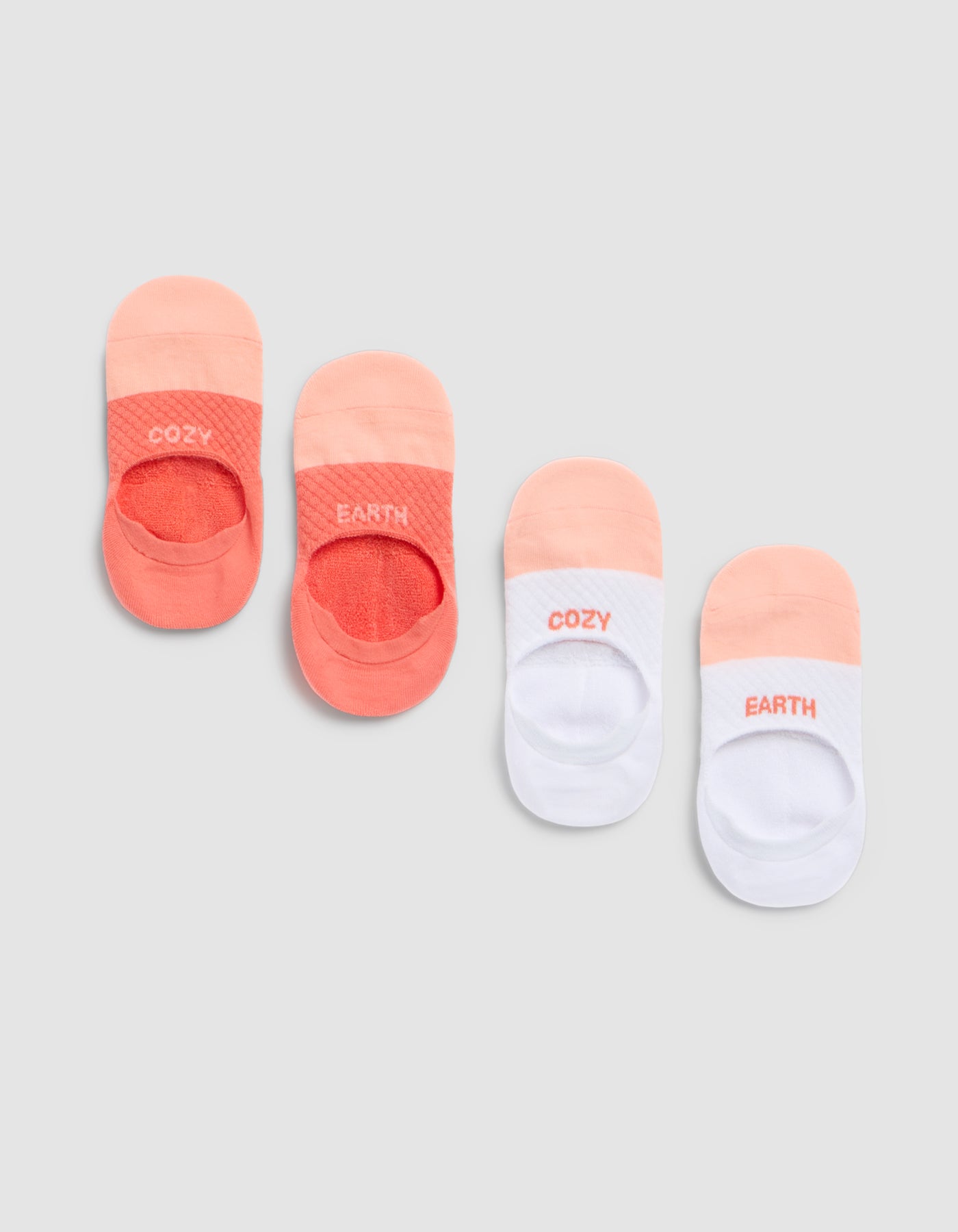 Four pairs of Cozy Earth Essential No-Show Socks are arranged side by side—two in coral tones and two in white with coral accents. Each sock features "COZY" or "EARTH" near the toes. 