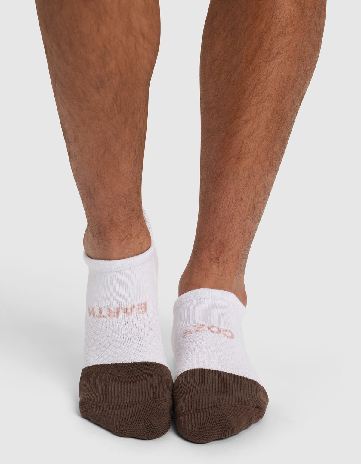 A person wears mismatched white Essential Ankle Socks by Cozy Earth with brown toes; one reads "EARTH" and the other "COZY" in light pink. They stand against a plain white background.