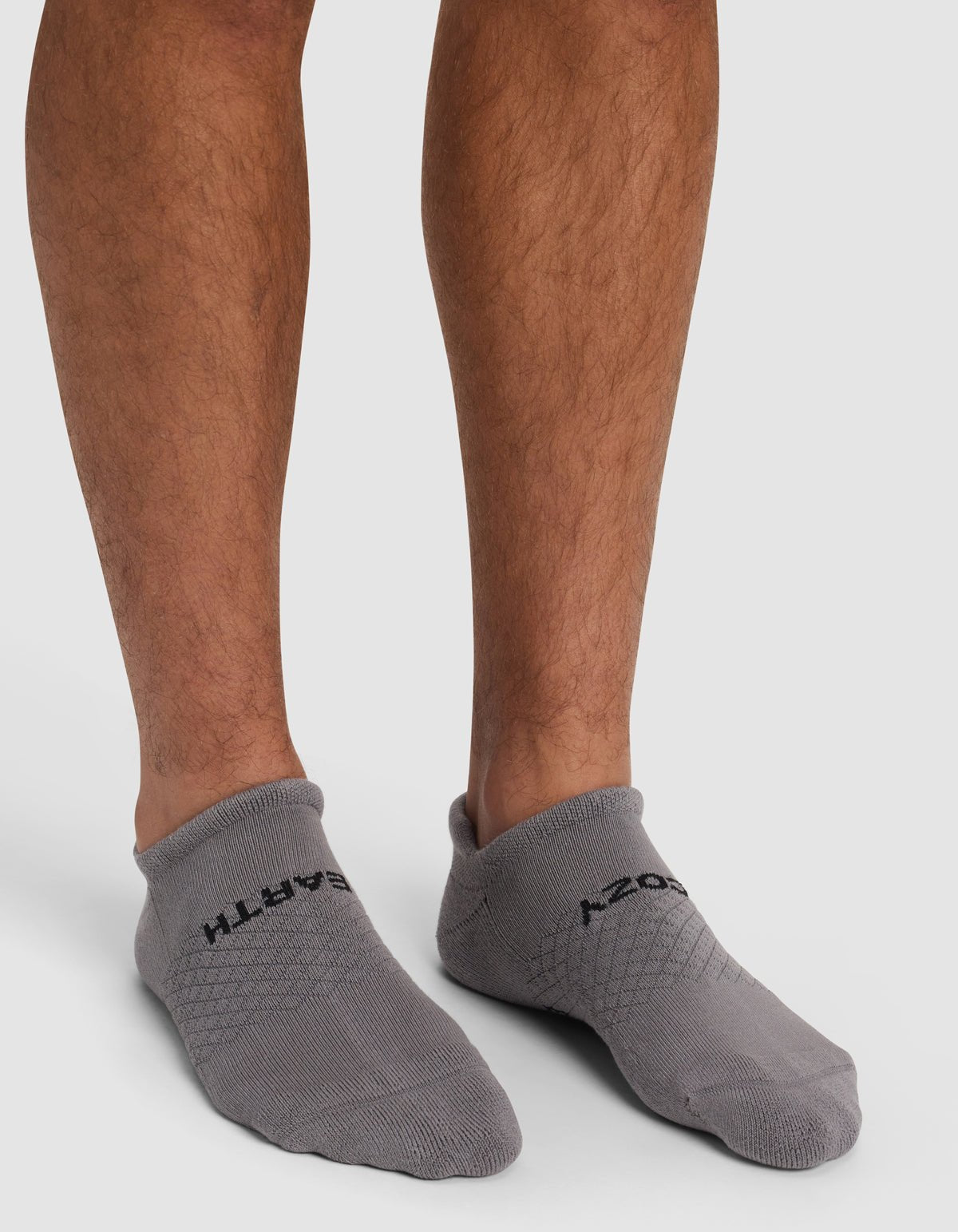 A person models Cozy Earth’s Essential Ankle Sock in gray with black text, standing on a white surface and showing only their lower legs and feet.