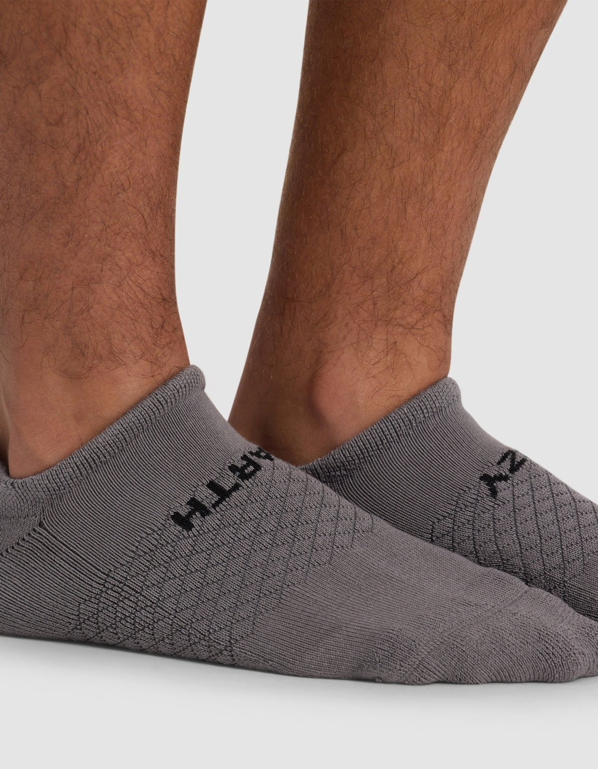 A close-up of a person's lower legs wearing Cozy Earth's Essential Ankle Sock in light gray with a subtle pattern and black text, standing on a light gray surface.