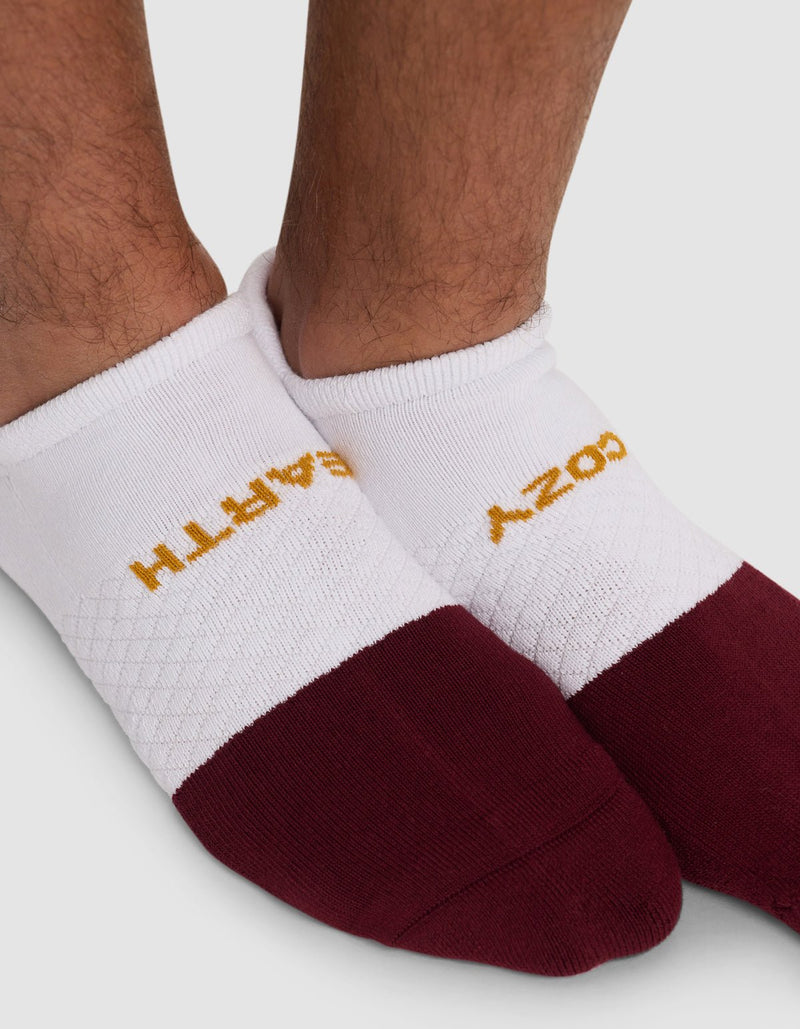 Close-up of a person's feet wearing Cozy Earth Essential Ankle Socks in white and maroon with yellow text on top, partially visible, against a plain light gray background.