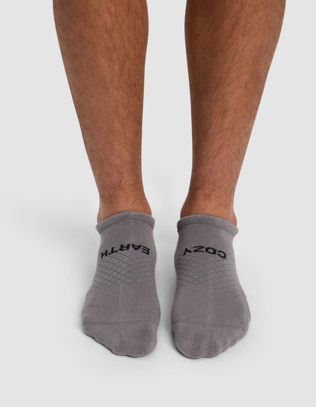 A person, with only their lower legs and feet visible, stands on a white background wearing Cozy Earth Essential Ankle Socks in gray. The left sock reads "EARTH" and the right reads "COZY.