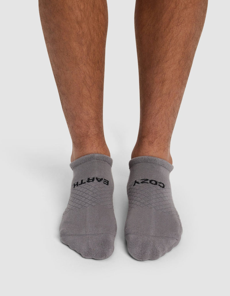 A person, with only their lower legs and feet visible, stands on a white background wearing Cozy Earth Essential Ankle Socks in gray. The left sock reads "EARTH" and the right reads "COZY.