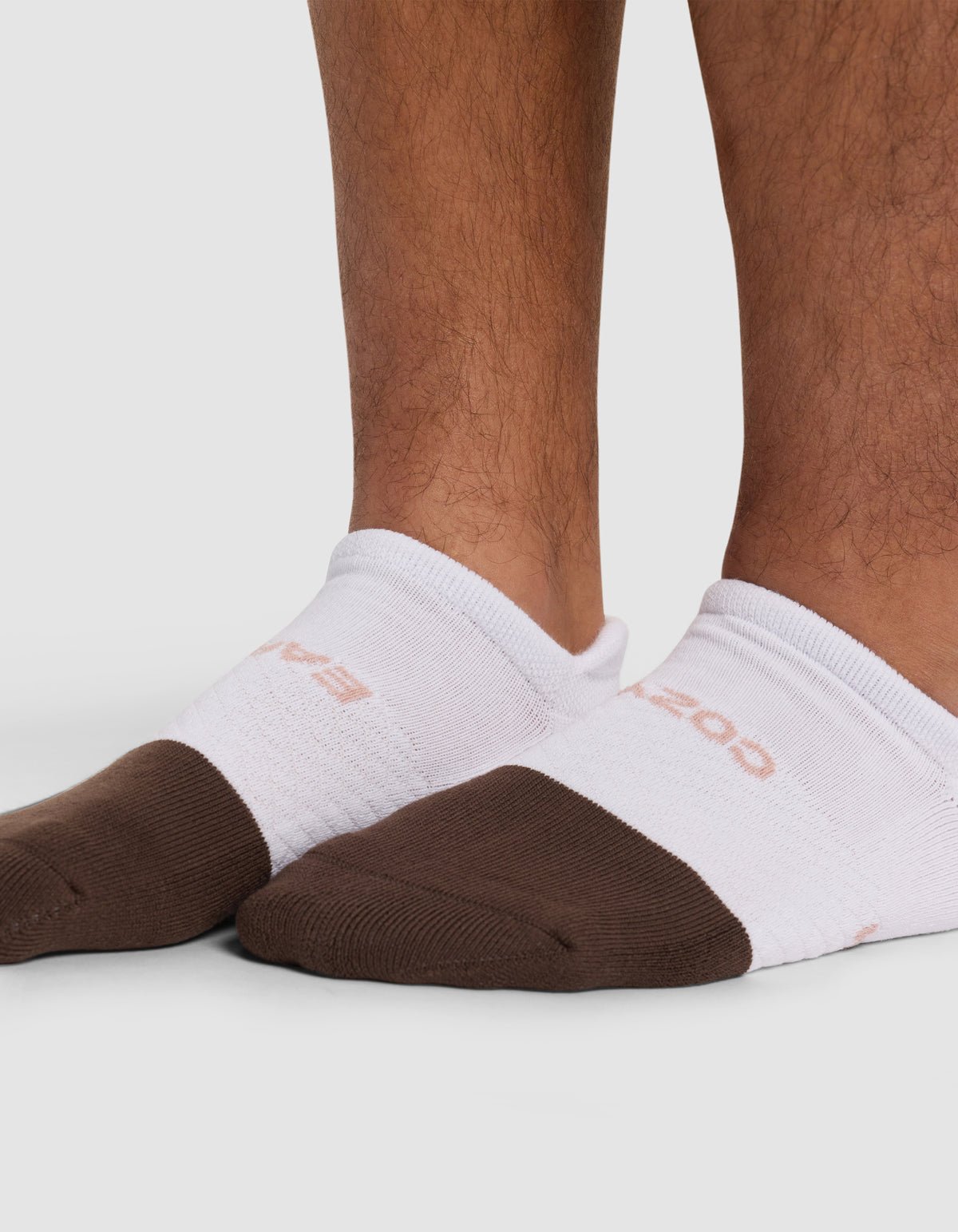 Close-up of a person’s lower legs wearing Cozy Earth Essential Ankle Socks in white with brown toes, standing on a white background.