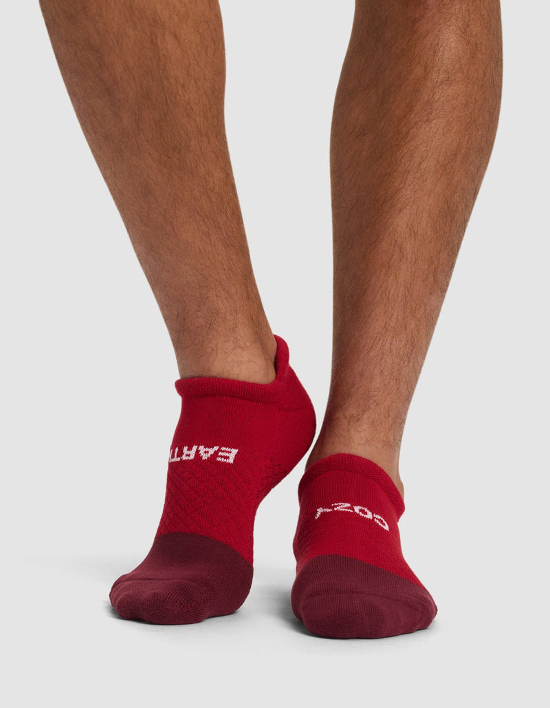 A person wearing Cozy Earth Essential Ankle Socks in red, featuring "EARTH" on the left and "GOOD" on the right at the top, stands on a white surface. Only their lower legs and feet are visible.