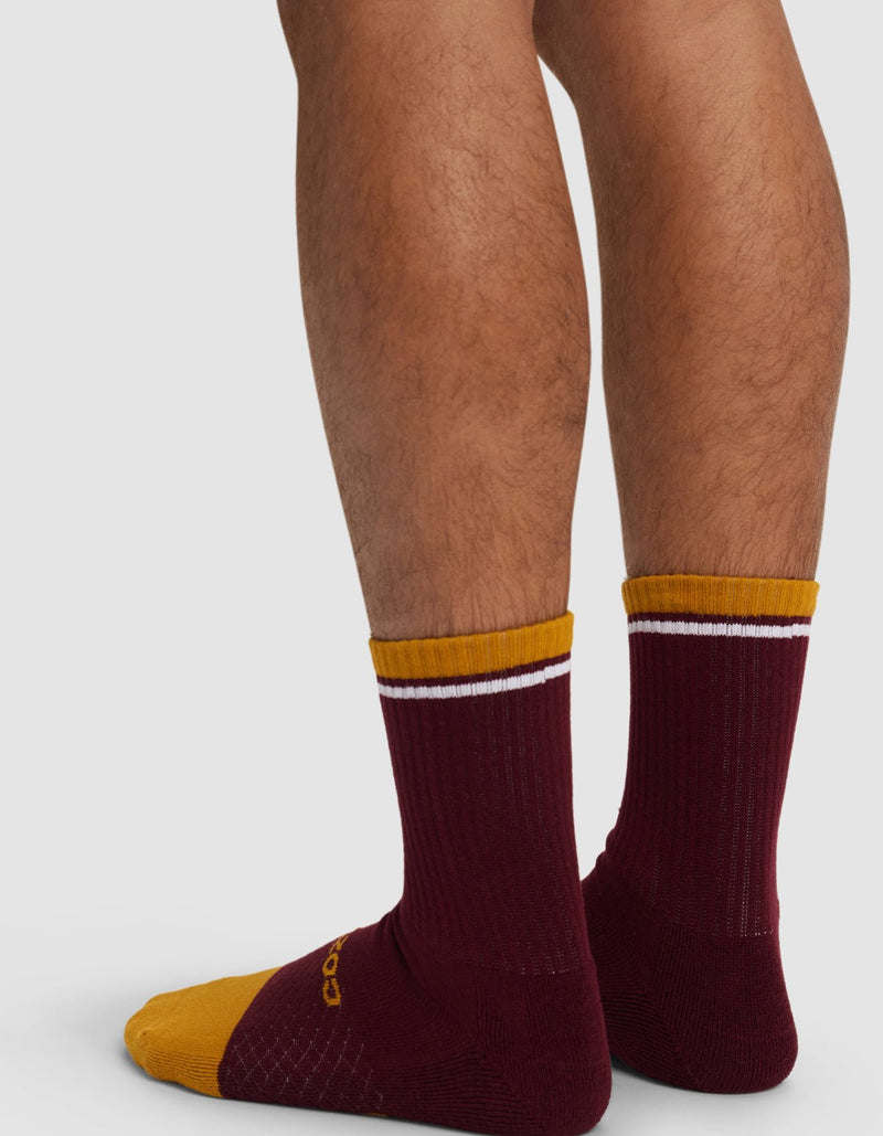 A person is wearing Cozy Earth Essential Calf Socks in maroon with yellow toes, yellow cuffs, and a white stripe near the top. Only their lower legs and feet are visible as they stand on a light surface.