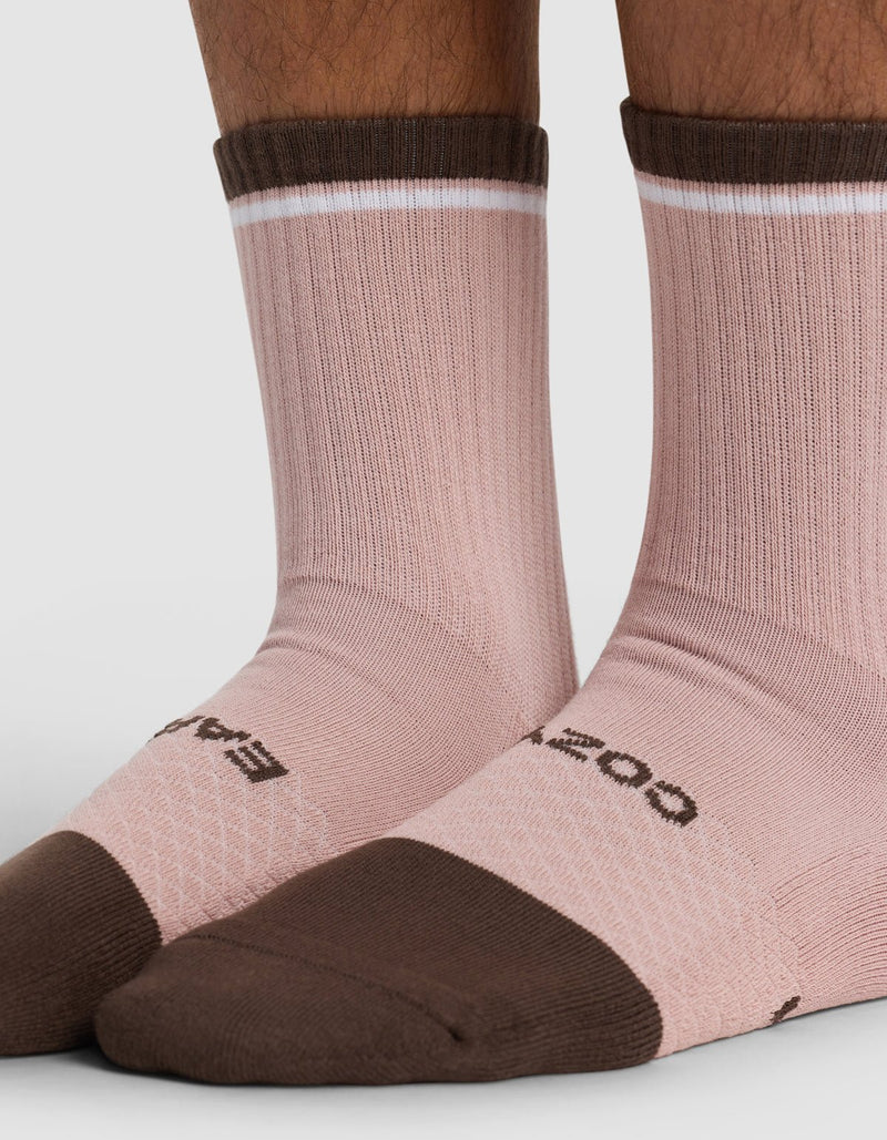 Close-up of a person's lower legs and feet wearing Cozy Earth Essential Calf Socks in light pink with dark brown toes, heels, cuffs, and a thin white stripe near the cuff. The brand name is partially visible on the socks.