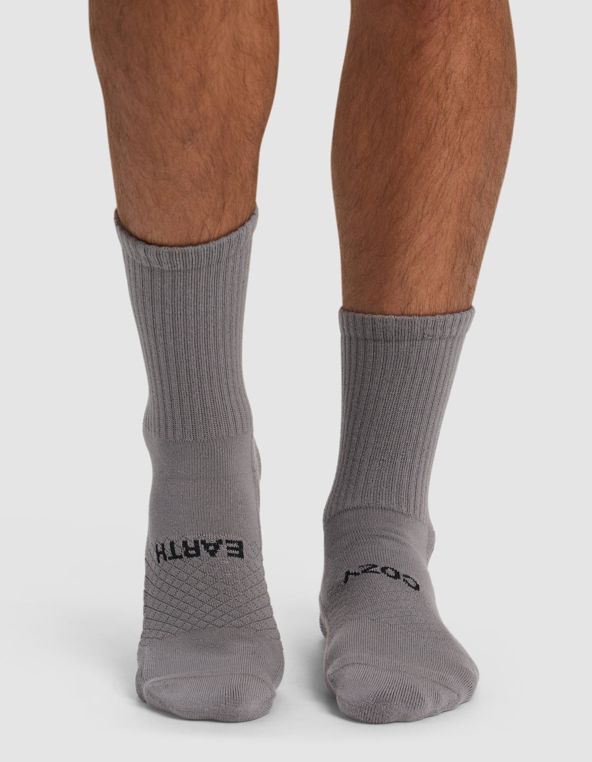 Someone stands on a light surface wearing Cozy Earth Essential Calf Socks in gray, featuring “EARTH” and “COSY” in black on the tops.