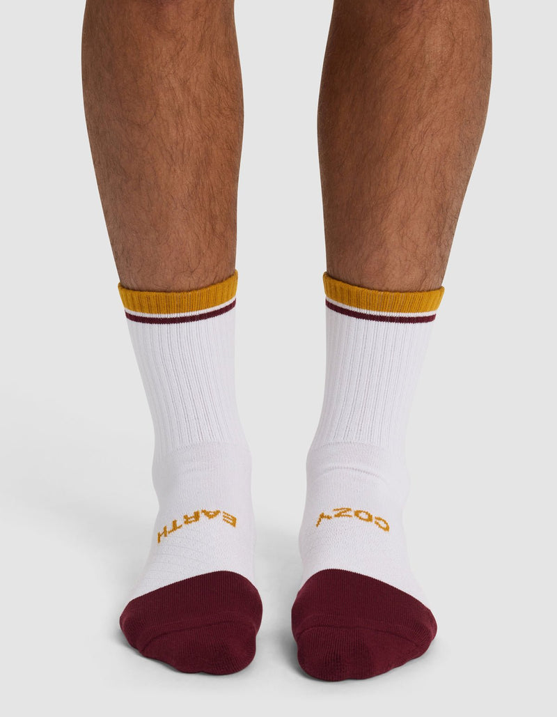 A pair of legs from the knees down wears Cozy Earth Essential Calf Socks—white with maroon toes and gold trim. "EARTH" in gold is on the left sock, "COSY" in gold on the right.