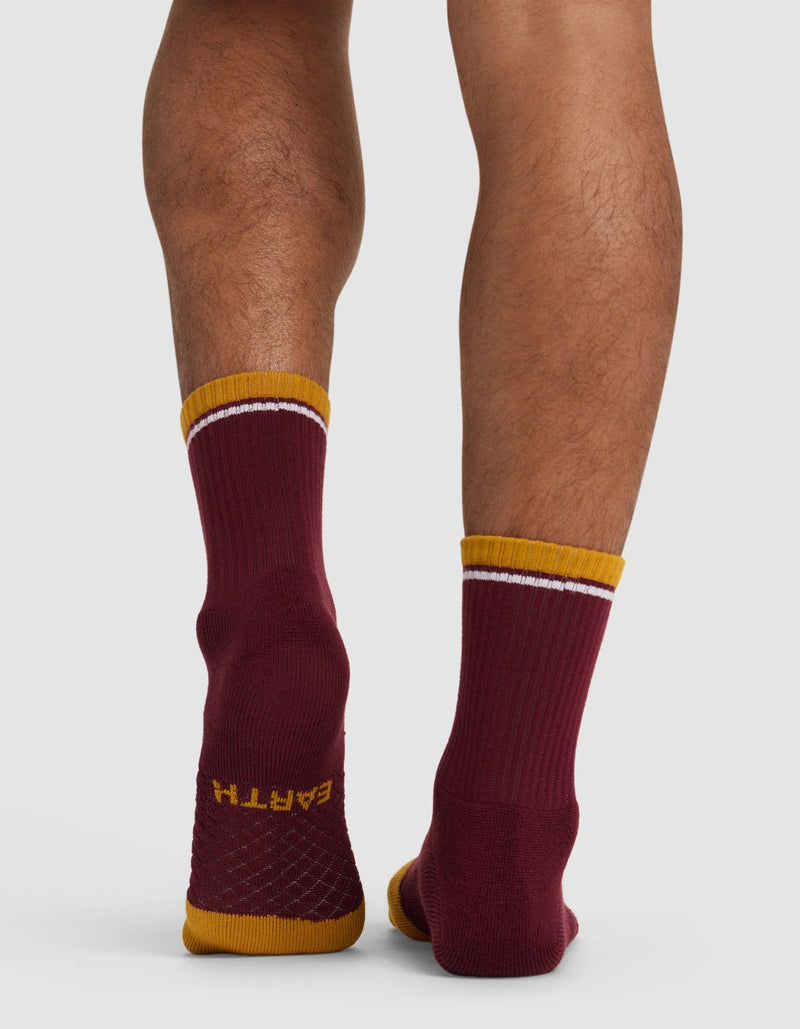 Back view of a person wearing Cozy Earth's Essential Calf Sock—mismatched maroon socks with gold toes and cuffs, white stripes near the top, and "EARTH" on one sole. The person stands on a light background.