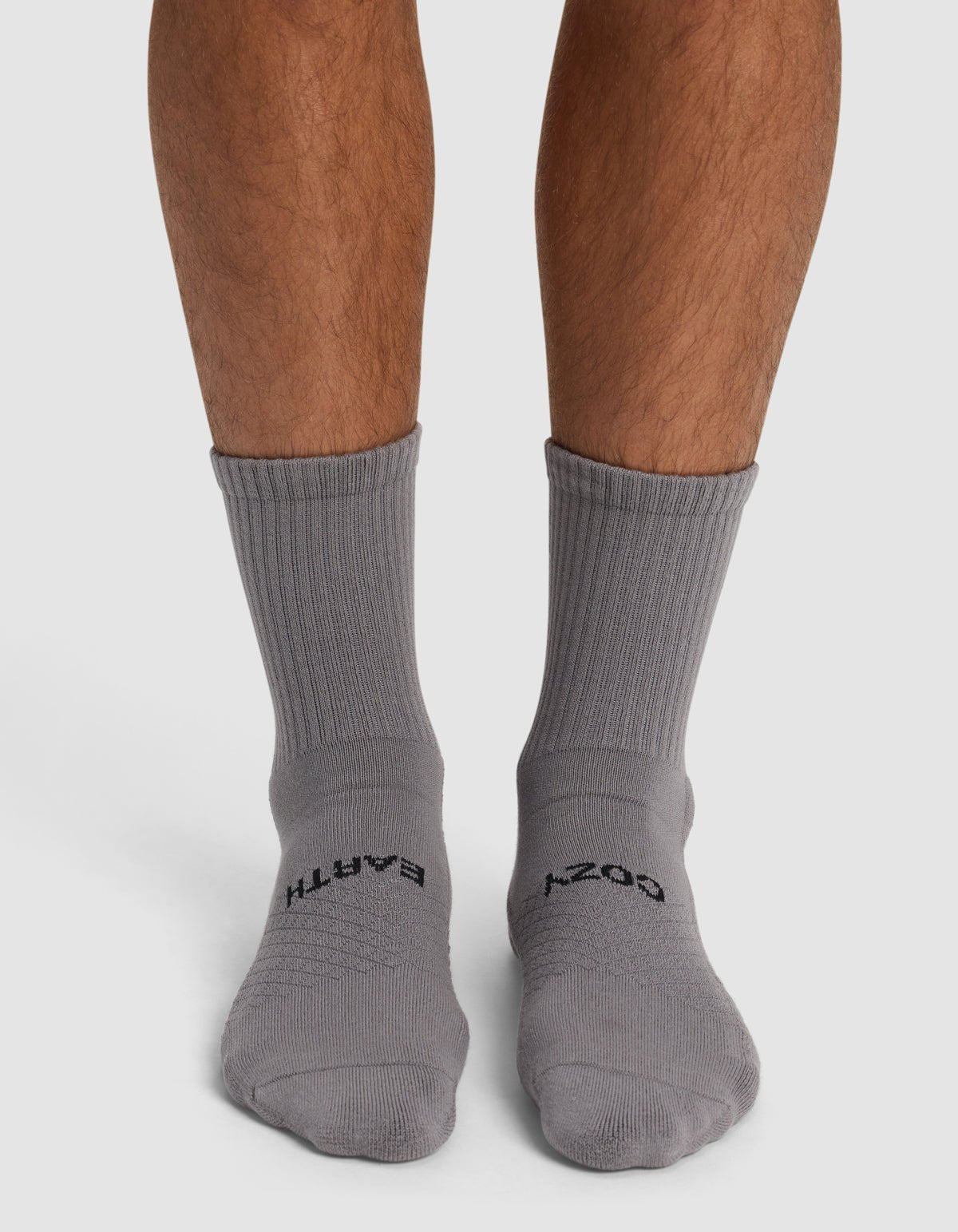 A person stands on a white surface wearing Cozy Earth Essential Calf Socks in gray—the left sock reads "EARTH" and the right sock reads "COZY" in black text.