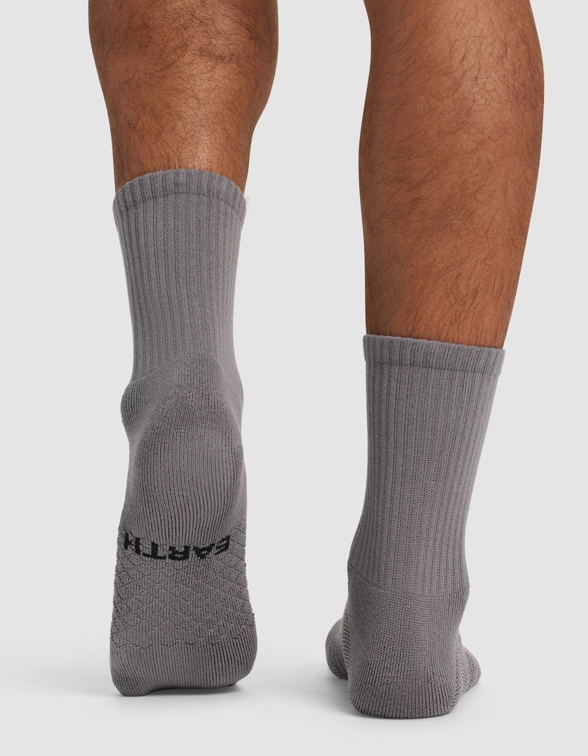 A person stands on a white surface wearing Cozy Earth Essential Calf Socks in gray. The back of the socks and lower legs are visible, with "EARTH" in black text on the sole of the left sock.