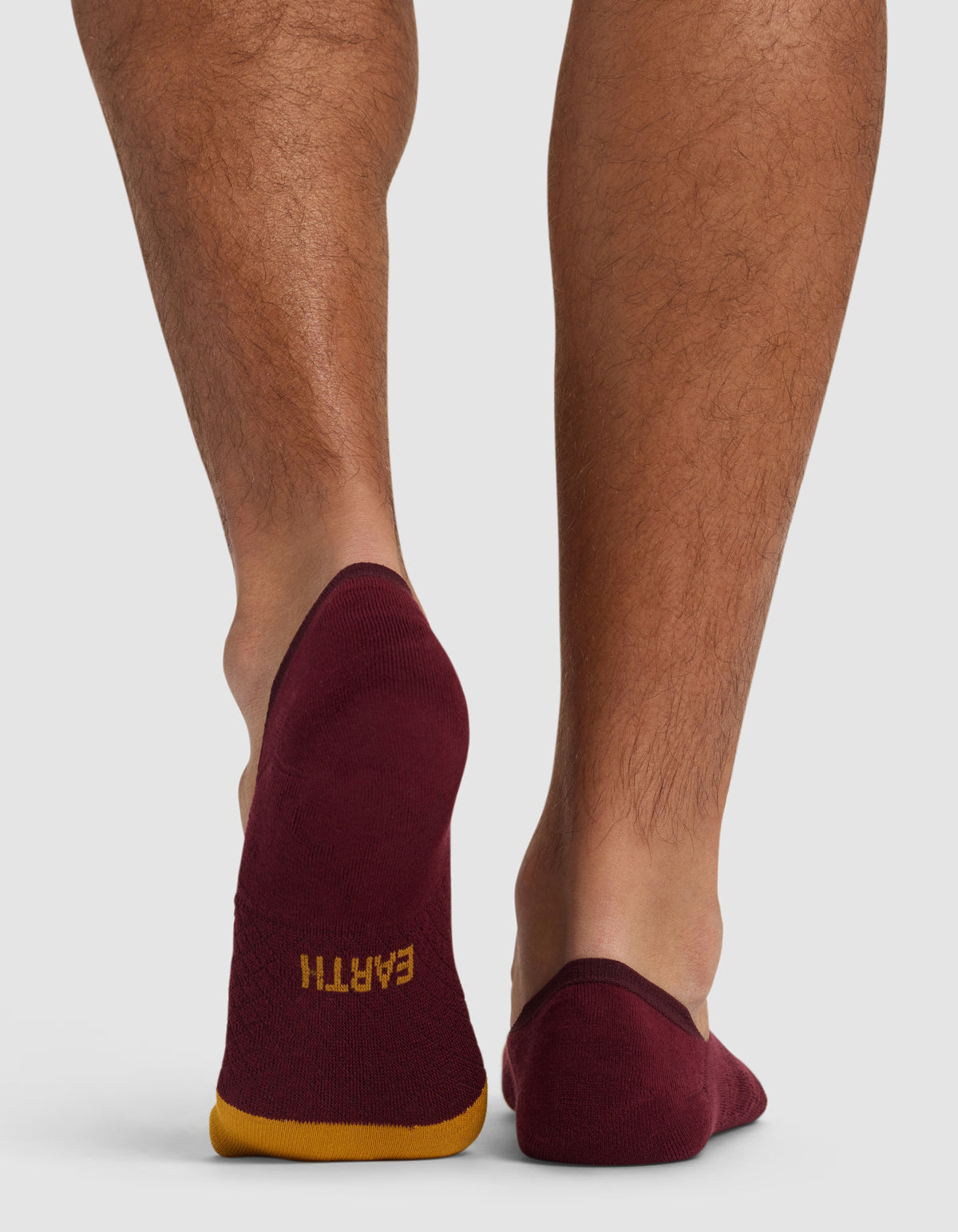 A person's lower legs and feet are shown from behind, wearing Cozy Earth's maroon Essential No-Show Socks with yellow trim and “EARTH” printed upside down on the left heel.