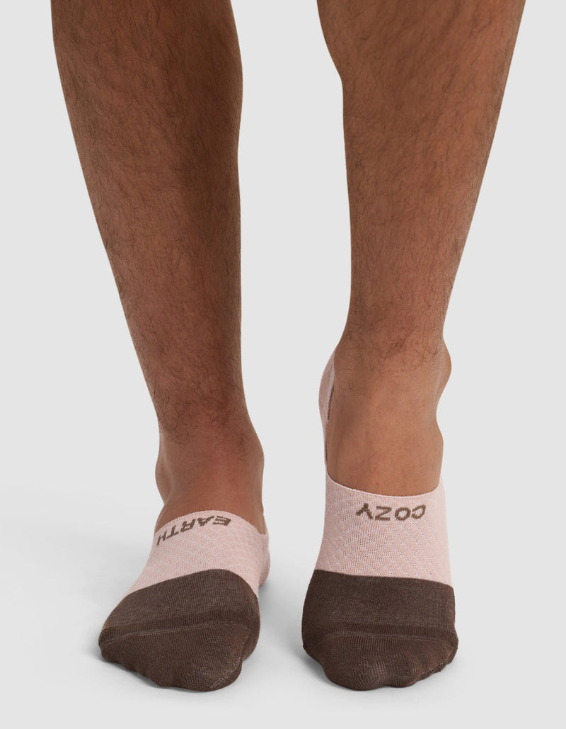 A person wearing Cozy Earth's Essential No-Show Socks in pink and brown, featuring "EARTH" on the left sock and "COZY" on the right, stands against a white background.