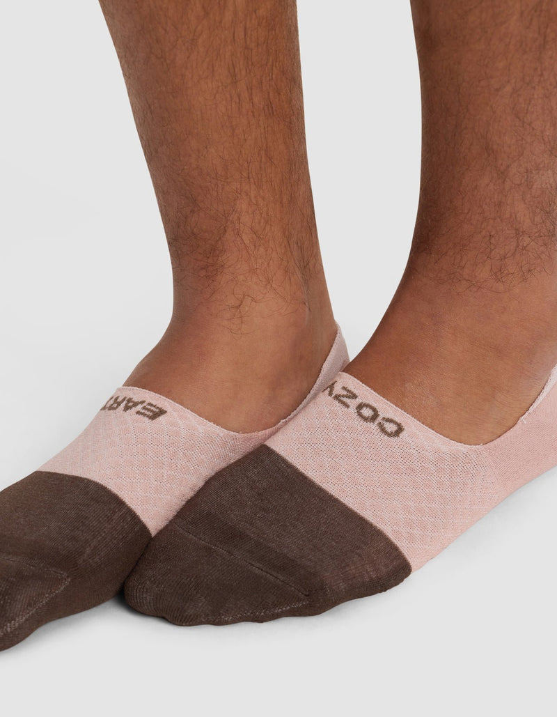 Close-up of a person's lower legs and feet wearing Cozy Earth Essential No-Show Socks in pink and brown with text near the ankle, standing on a light surface.