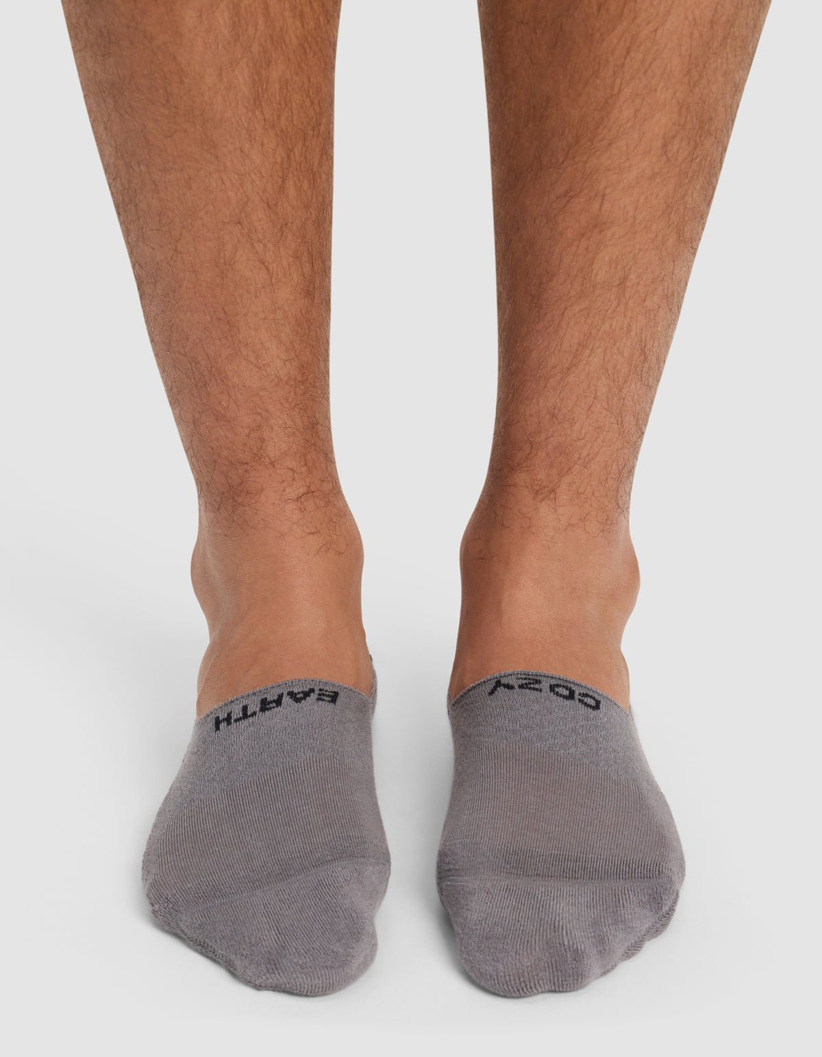 A pair of feet in Cozy Earth Essential No-Show Socks in gray, featuring "GO EARTH" near the top, standing on a white surface.