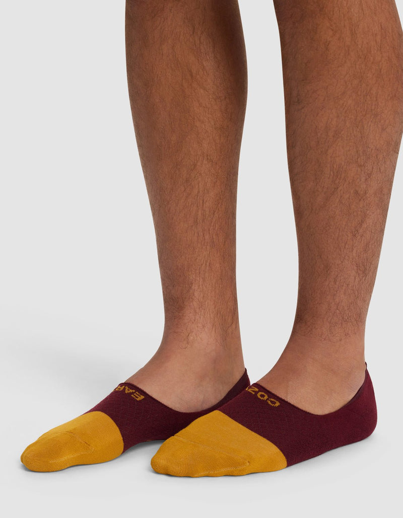 A person wears Cozy Earth Essential No-Show Socks in maroon and yellow, standing on a white surface with only their lower legs and feet visible.