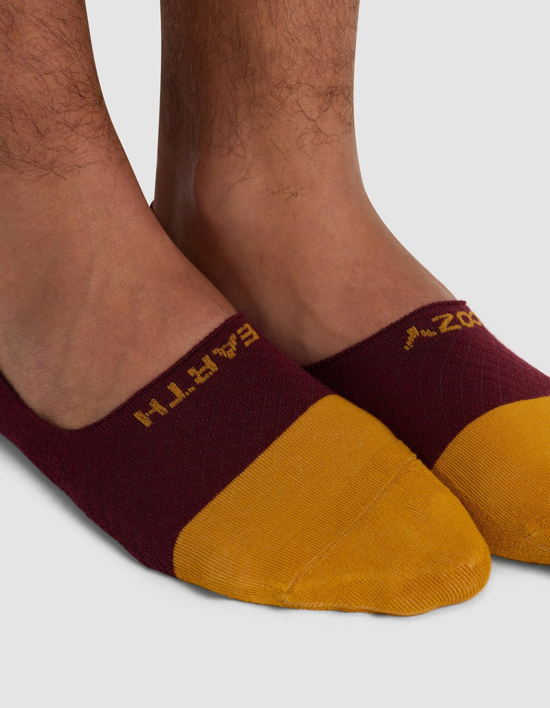 Close-up of a person's feet in Cozy Earth Essential No-Show Socks, maroon and yellow with yellow text by the opening, standing on a light gray surface.