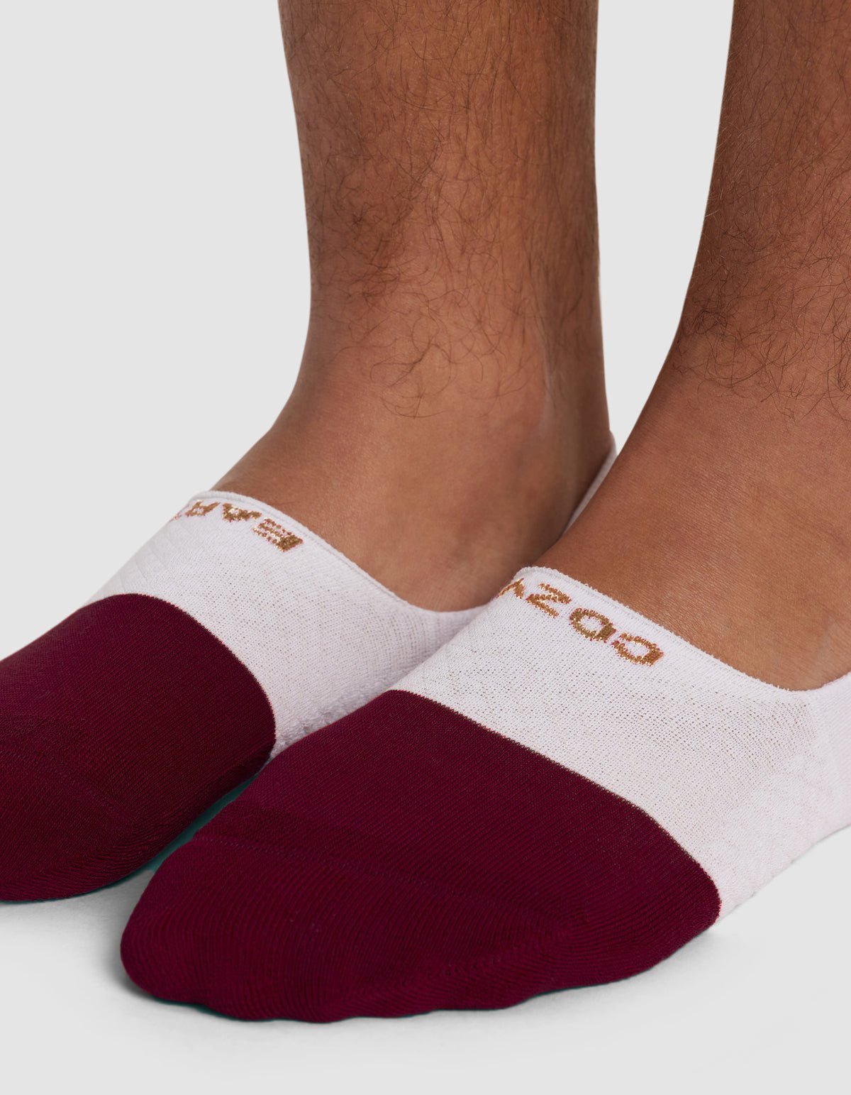 A close-up of feet wearing Cozy Earth Essential No-Show Socks, featuring maroon toes, white midsections, and a tan logo near the ankle, set against a plain light background.