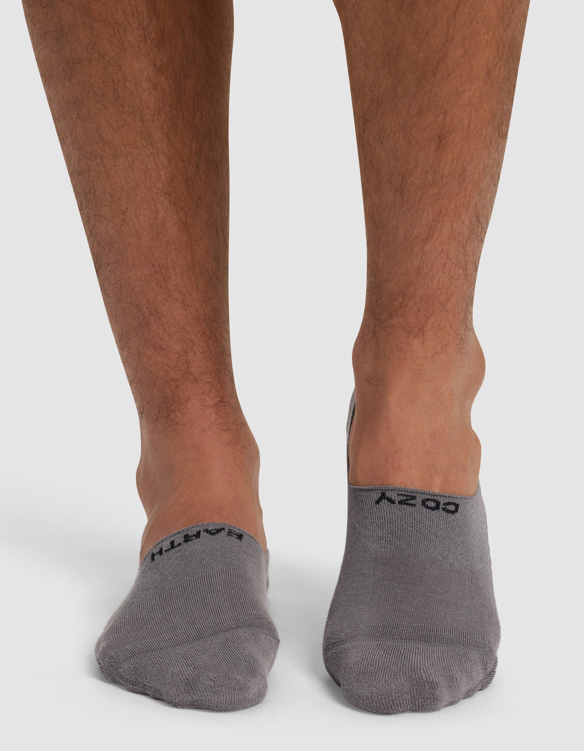 A close-up shows a person's lower legs and feet on a white surface, wearing Cozy Earth's Essential No-Show Socks in gray, each featuring black text on top.