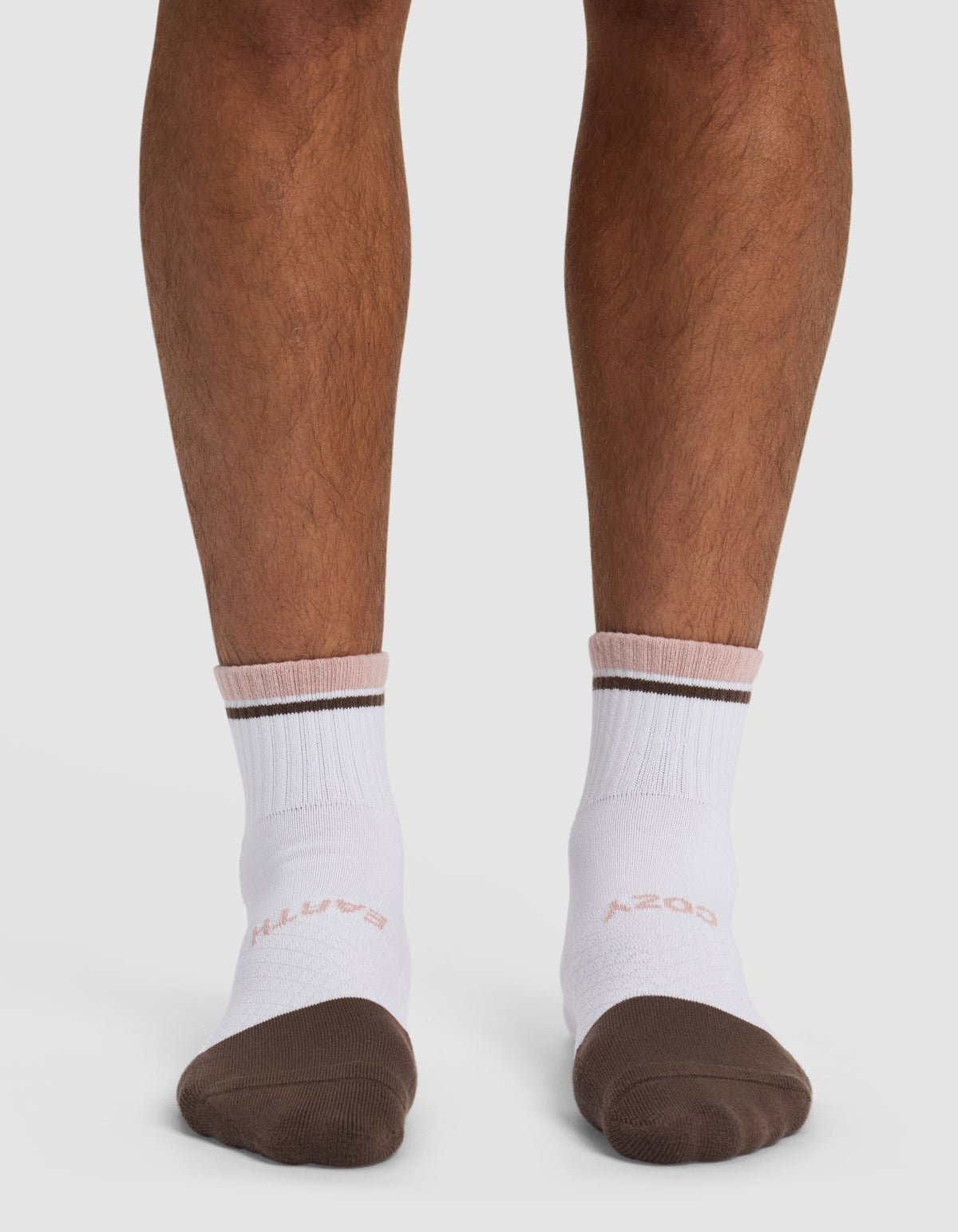 A person stands on a white surface wearing Cozy Earth Essential Quarter Socks—white ankle socks with brown toes, a brown stripe near the top, and "COZY" on one sock and "EARTH" on the other.