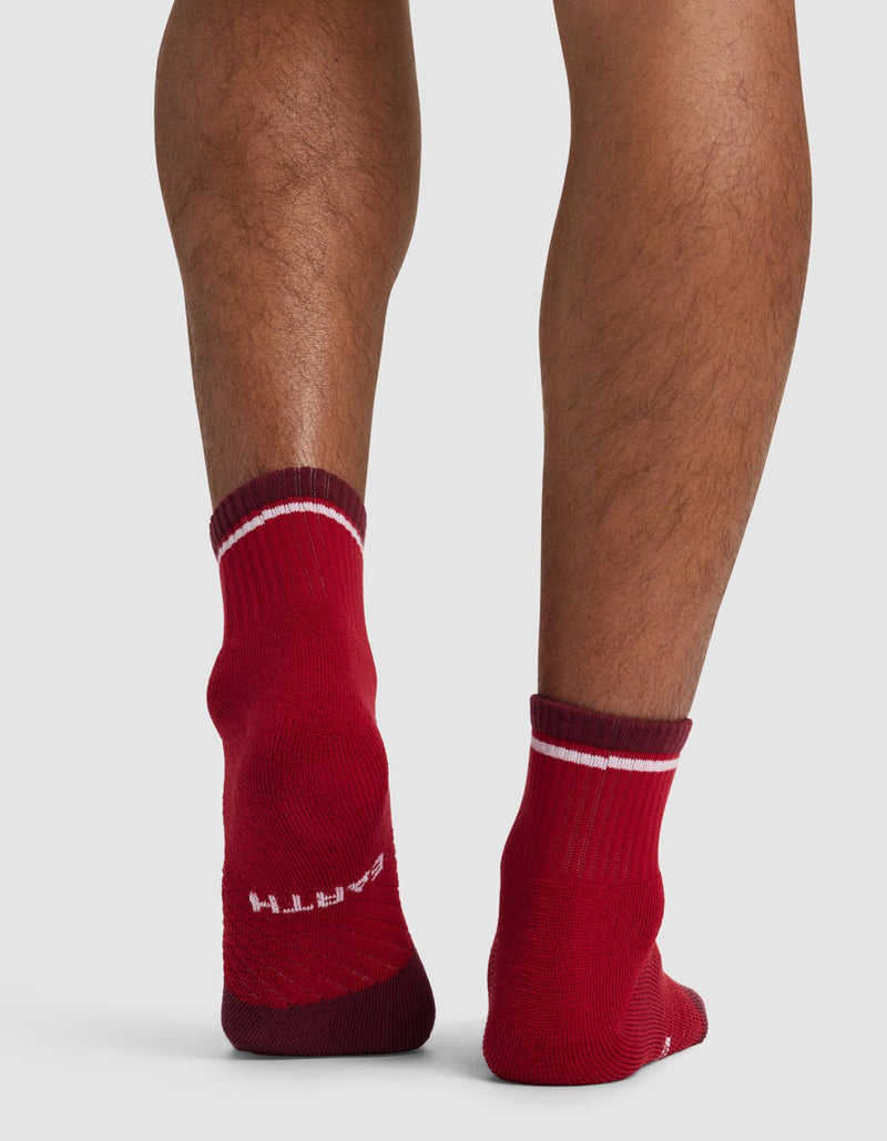 Someone stands on a light-colored surface, showing the back of their legs in Cozy Earth Essential Quarter Socks—red with white stripes. The word "EARTH" is visible on the heel of the left sock.