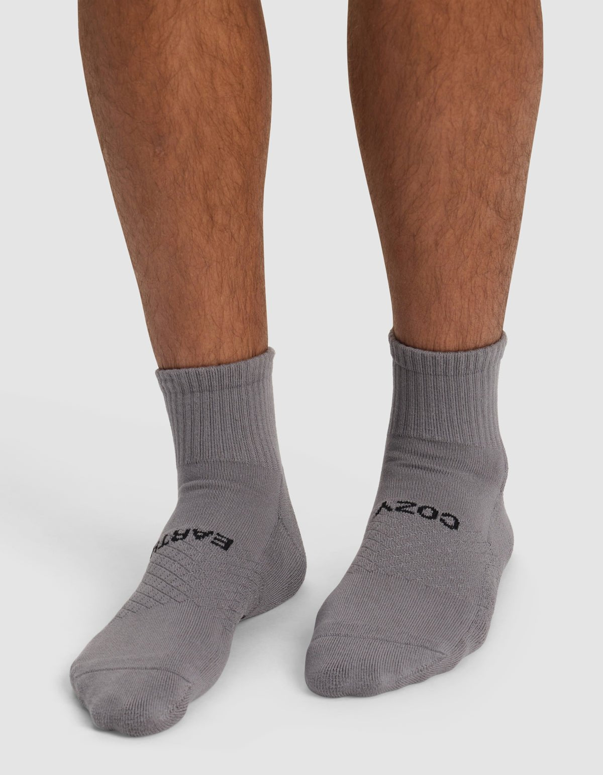 A person wearing Cozy Earth Essential Quarter Socks in gray with black text, standing on a white surface. The socks cover the ankles and part of the lower legs.
