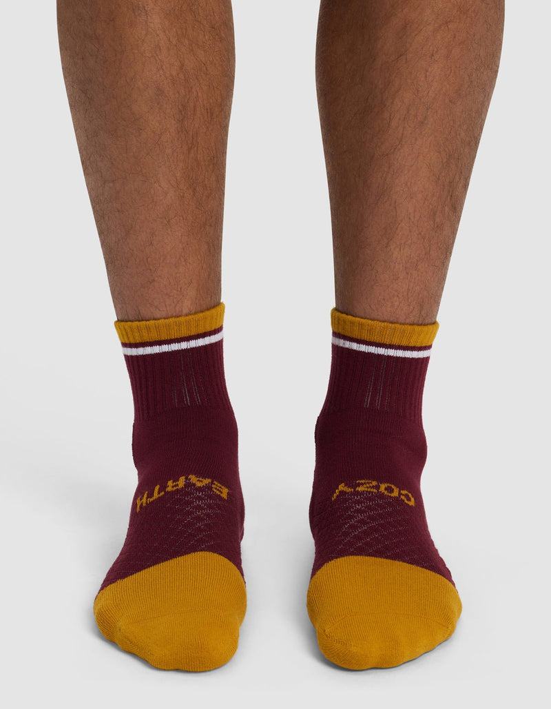 A person wears Cozy Earth Essential Quarter Socks in maroon and yellow with white top stripes, yellow toes, and text on the foot. Their lower legs are visible against a plain background.