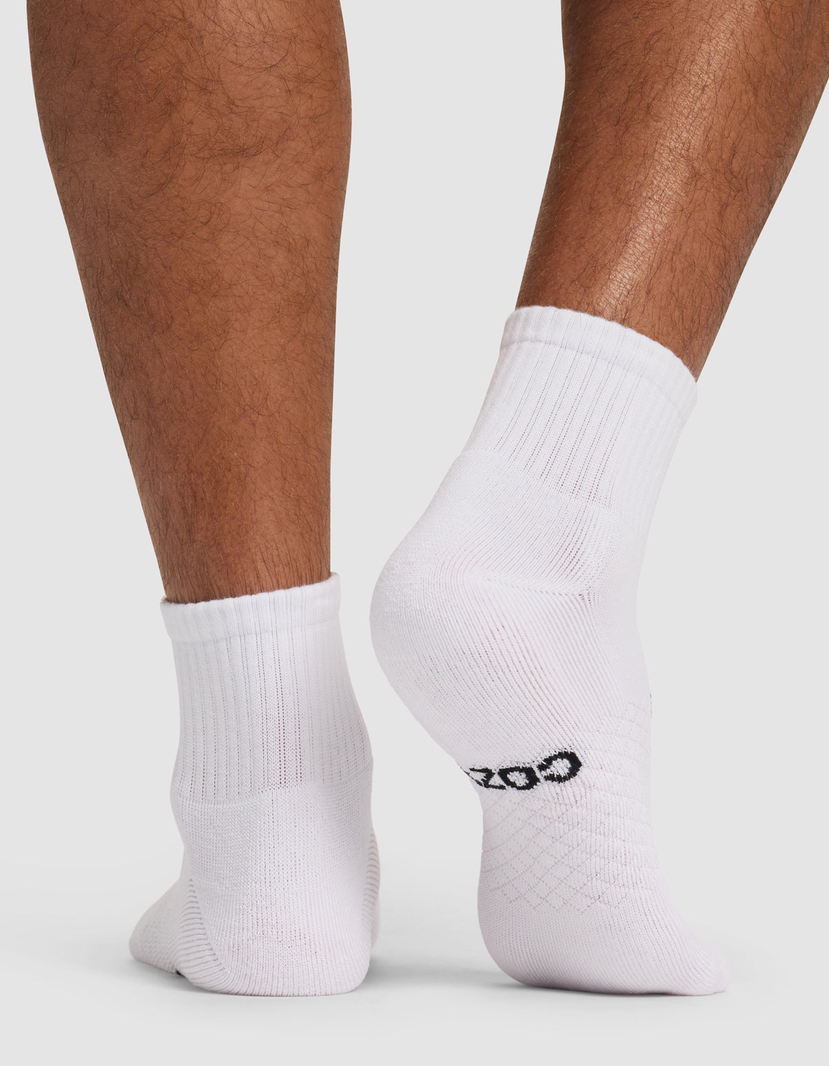 Close-up of a person’s lower legs and feet wearing Cozy Earth’s Men’s Essential Quarter Socks in white. The person is standing on a white background, with one foot slightly raised to display the sock’s pattern and a small black logo on the sole. Shown as part of the Men’s Essential Quarter Sock 4-Pack – Last Chance.