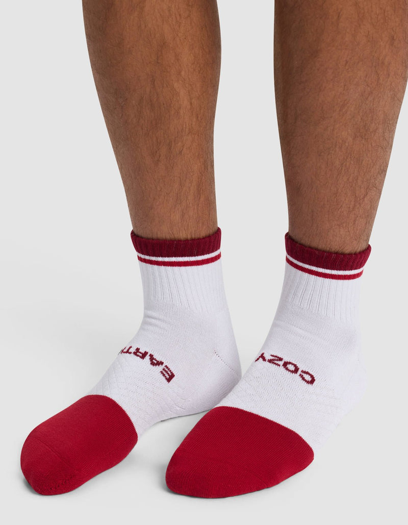 A person wears Cozy Earth Essential Quarter Socks—white with red toes, heels, and cuffs. The left sock reads "EARTH" in red, the right reads "COZY." They are standing on a light surface.