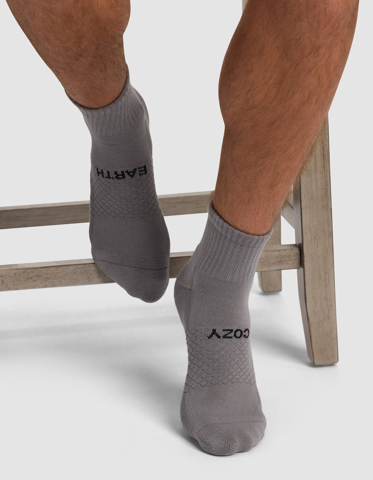 A person sits cross-legged on a wooden stool wearing Cozy Earth Essential Quarter Socks in light gray, featuring "EARTH" and "COZY" in black lettering.