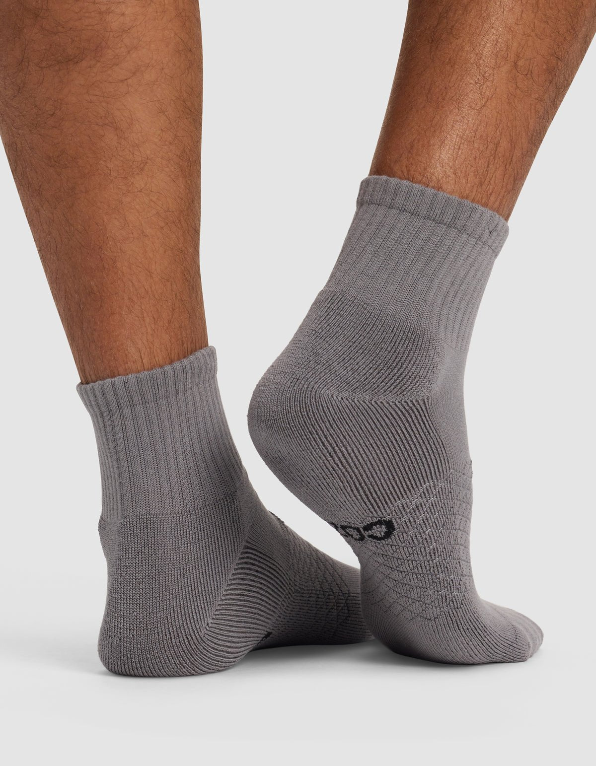Close-up of a person's lower legs wearing Cozy Earth Essential Quarter Socks in light gray with ribbed cuffs, standing on a white surface. A small black logo appears near the heel of one sock.