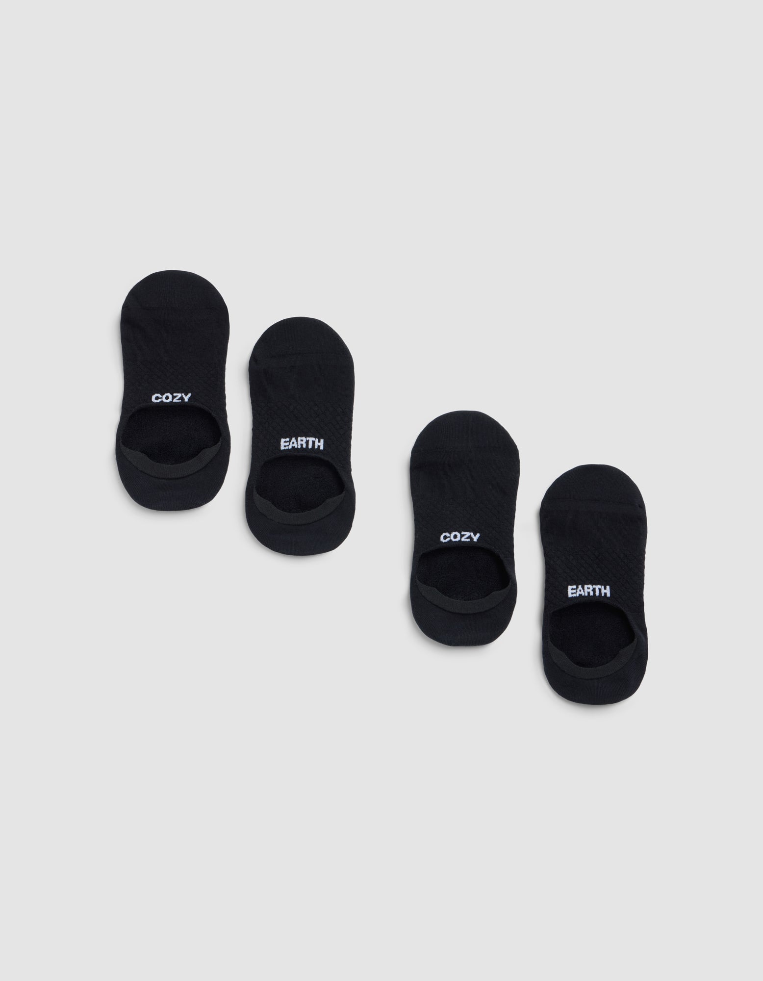 Two pairs of black Essential No-Show Socks by Cozy Earth are displayed on a light gray background. One pair features "COZY" in white lettering, and the other pair displays "EARTH" in white. 