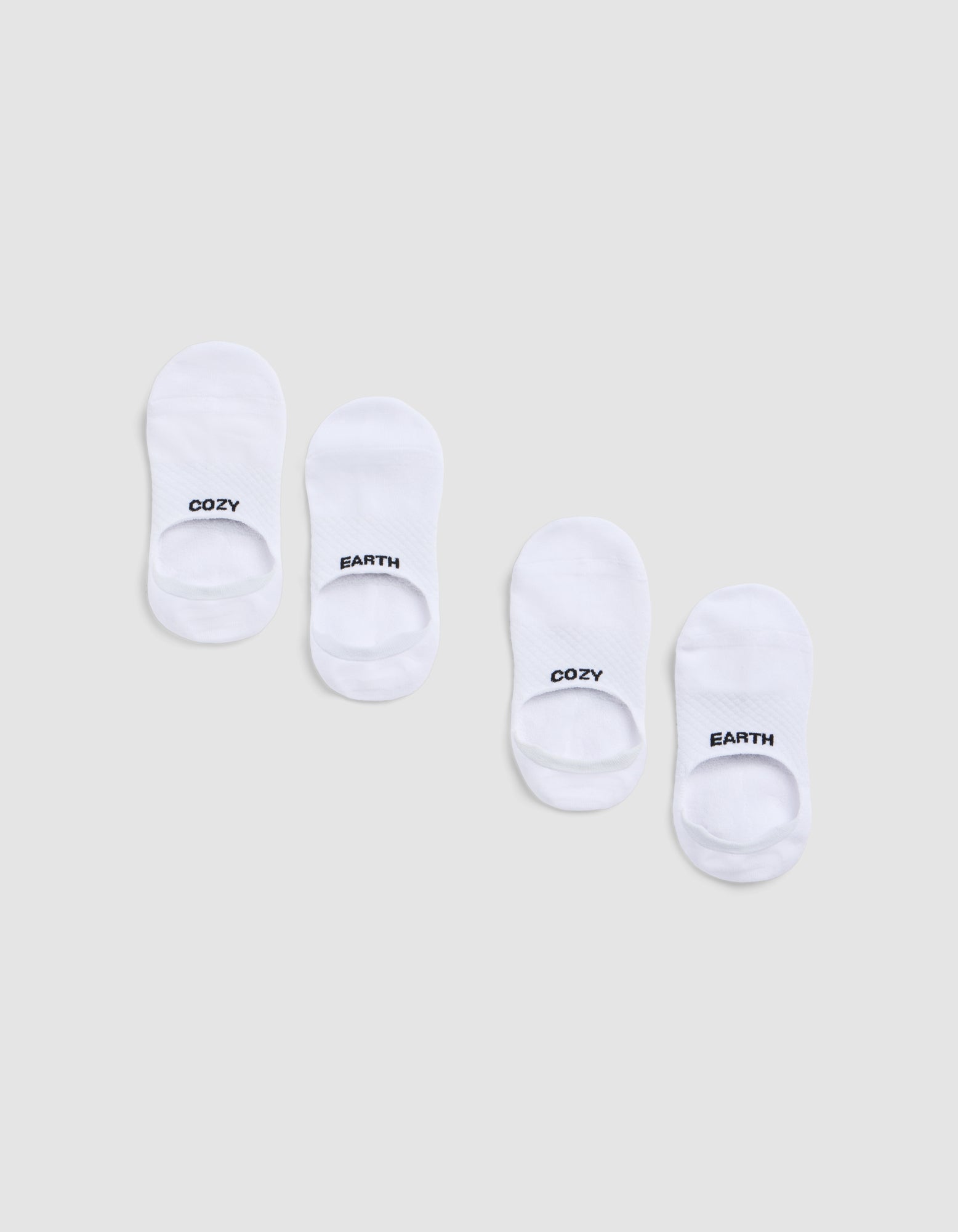 Two pairs of Cozy Earth Essential No-Show Socks are displayed on a light gray background. Each pair features "COZY" on one sock and "EARTH" on the other, both printed in black near the toes. |Color:White