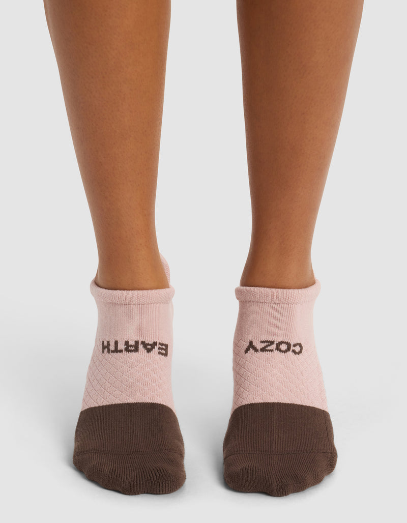 A person wears Cozy Earth Essential Ankle Socks in pink and brown; "EARTH" is written on the left sock and "COZY" on the right, both in black letters. The background is plain white.